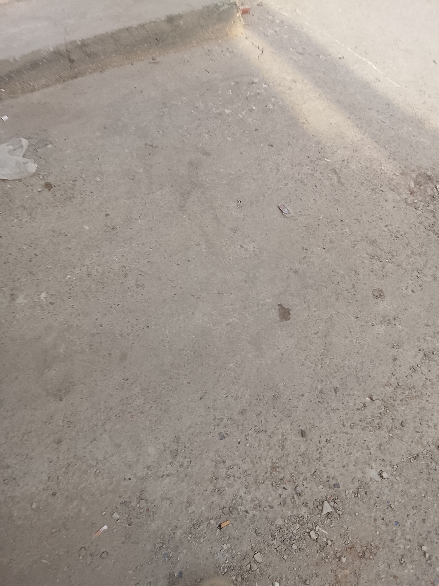 The image depicts a section of weathered concrete ground in Gulgasht Colony, Pakistan. The surface is uneven, with a mix of fine and coarse aggregates. There is debris scattered across the ground, including a small piece of white plastic, small rocks, and what appears to be a small piece of brown paper. A concrete curb can be seen in the upper left corner of the image. Sunlight casts shadows across the scene, creating highlights and darker areas on the surface.