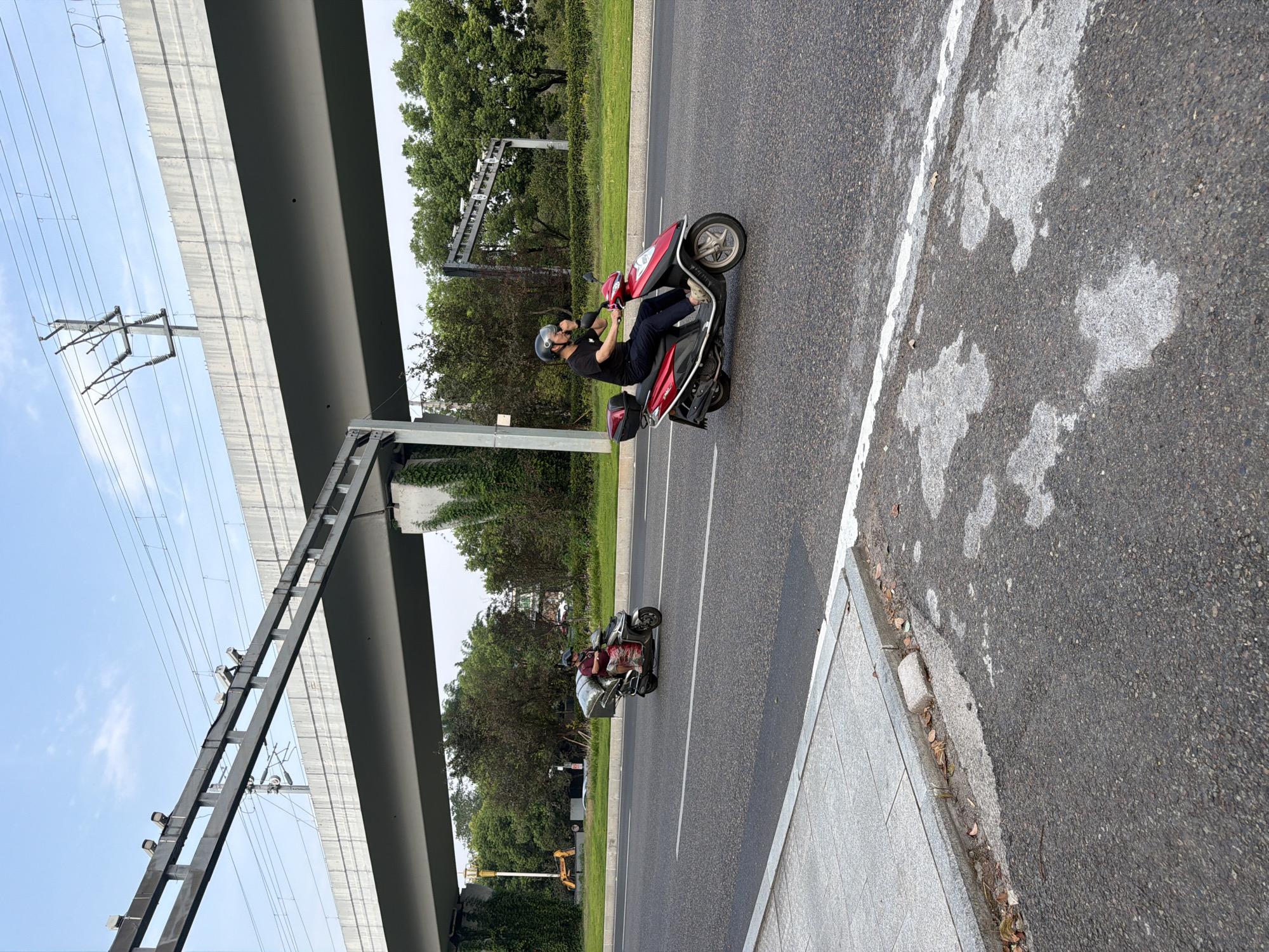 In Jiangjin, China, two individuals ride scooters on a paved road beneath an elevated concrete structure, likely a highway or railway overpass. The structure is supported by thick concrete pillars, and a metal framework with what appear to be traffic cameras is mounted above it. Electrical wires and poles are visible in the background against a sky with scattered clouds.
On the road, a person wearing a dark shirt and helmet rides a red and black scooter, looking towards the right. Behind them, another person in a maroon shirt and a helmet is on a gray scooter, carrying a large silver box on the back. They are also traveling in the same direction.
To the left of the road, a sidewalk with gray paving stones borders the asphalt. The environment is lush with green trees and vegetation, suggesting a suburban or semi-rural setting within the city. The road surface shows signs of wear, with some patches of lighter asphalt and white lane markings.