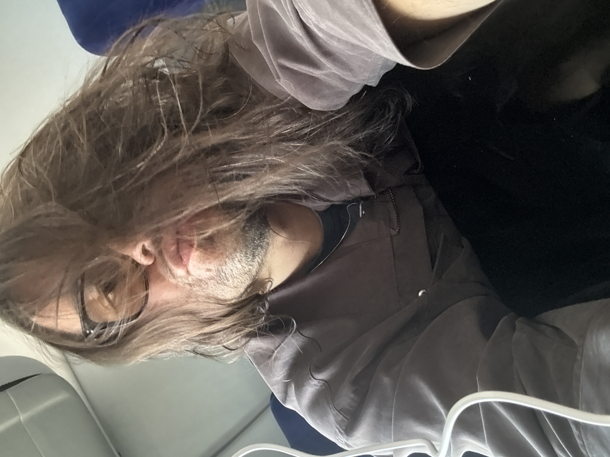 The media file displays a rotated, close-up selfie of a person, likely male, with long, disheveled brown hair falling over their face. The person wears black-framed glasses and has light stubble or a short beard. They are dressed in a light grey or taupe long-sleeved shirt or jacket, with a darker blue garment visible underneath.
The subject is positioned with their head slightly tilted, and their gaze is directed towards the camera, conveying a neutral or slightly pensive expression. The hair appears somewhat textured, possibly damp or styled to look messy. Details from the crops highlight the individual strands of hair, the texture of the stubble, and the folds in the fabric of the clothing. A small, metallic button or zipper pull is discernible on the shirt/jacket.
The setting appears to be an indoor environment. On the left side of the frame, a light grey structure, possibly a seatback, headrest, or part of an interior panel of a vehicle (like a plane, train, or bus), is visible. The background on the right is predominantly dark, suggesting limited light or an unlit area behind the subject. The overall lighting is subdued, with some highlights on the hair and face. There is no visible text in the image. The image itself does not contain visual cues directly indicating the specified location context of Valle de Chalco Solidaridad, Mexico; it only suggests an internal, possibly transportation-related, environment.