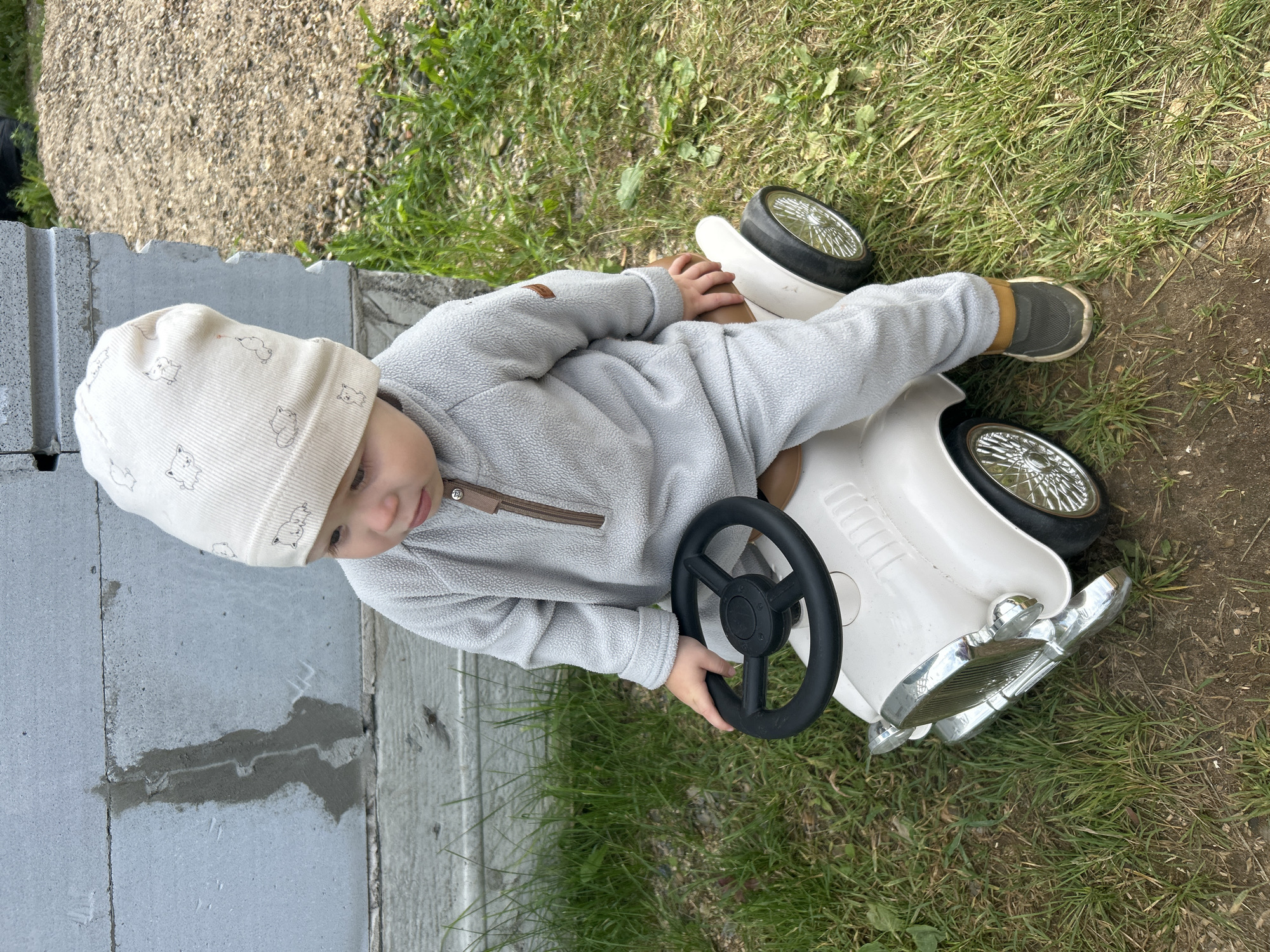 A young child, likely a toddler, is sitting on a white toy car outdoors. The child is wearing a light gray fleece jumpsuit with a zipper and a matching beanie hat with a pattern of cartoon animals. The child's right hand is on the black steering wheel of the car. The toy car has chrome accents on the front grill and headlights, and spoked wheels. The car is positioned on a grassy area. In the background, a wall constructed of grey concrete blocks is visible, with some patchy grey material on its surface. To the right of the wall, a pile of dirt and gravel is present, with green grass growing around it. The overall setting suggests a yard or an outdoor play area in a residential or possibly industrial setting. The location is identified as Chaikovsky, Russia.