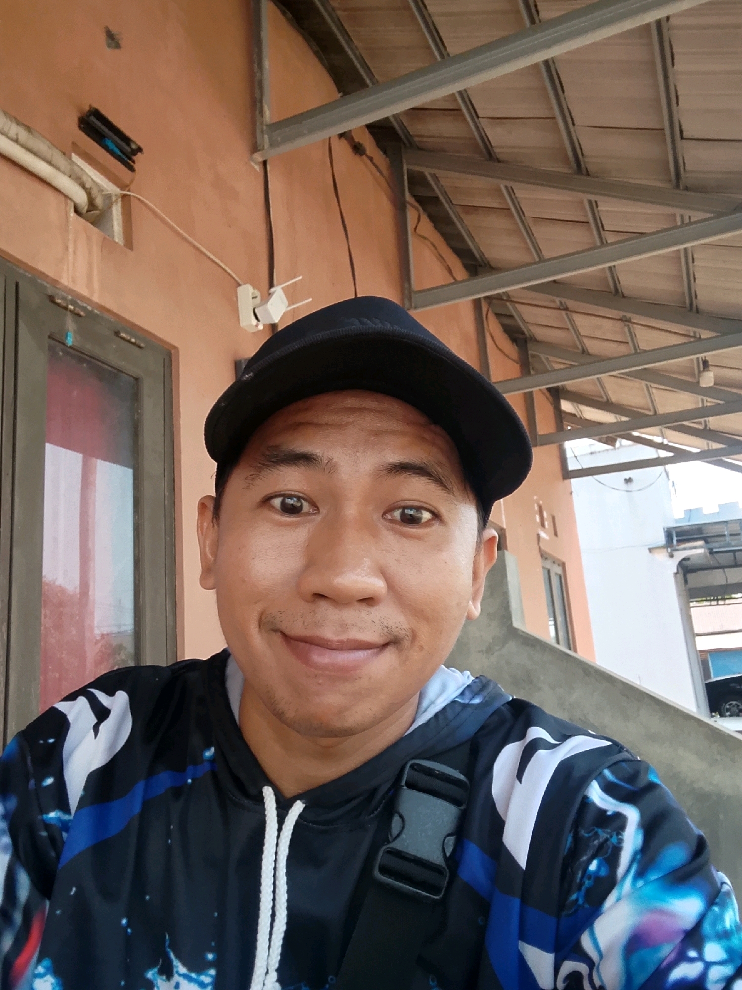 A medium shot selfie shows a man with a black baseball cap and a black, blue, and white patterned hoodie. He has brown skin, dark eyes, and a mustache. He is smiling at the camera. Behind him, a light brown wall is visible, with a window and a door on the left. A grey roof structure with wooden planks is visible overhead. The scene appears to be outdoors, possibly in Pelaihari, Indonesia, given the architectural style and materials.