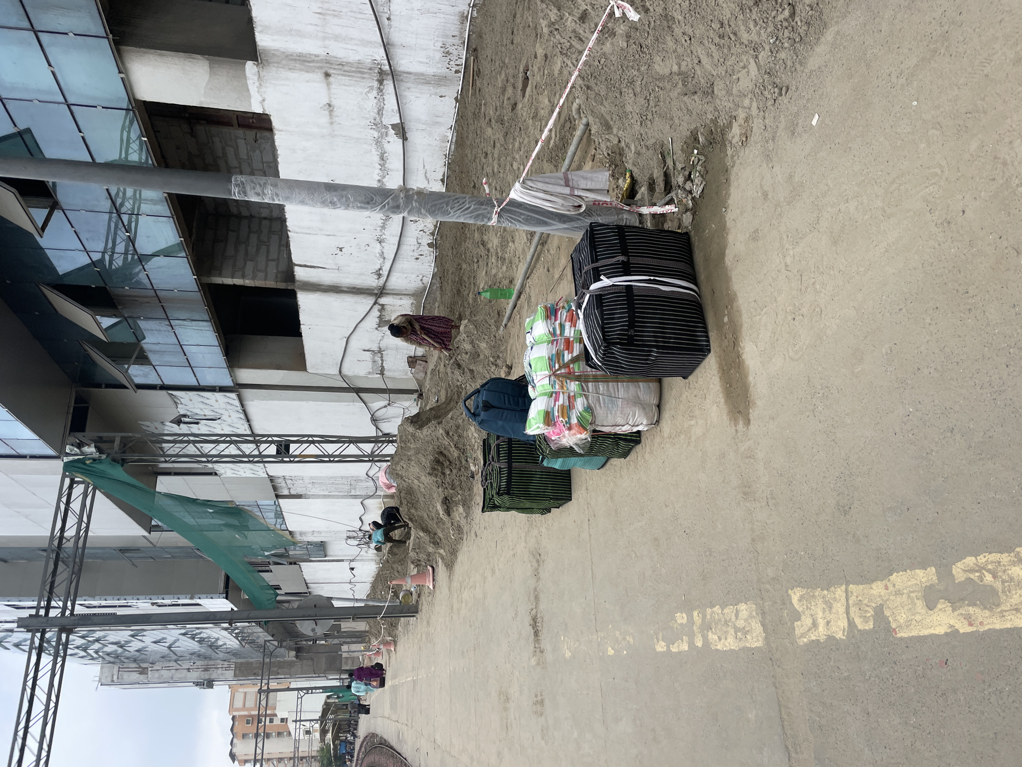 This image depicts a construction site adjacent to a train platform, likely in New Delhi, India, due to the architectural style and presence of train tracks. Several individuals are engaged in manual labor, shoveling dirt and sand into piles. The construction area is cordoned off with red and white tape, and a green mesh net is draped over some scaffolding, suggesting ongoing building work. A large, modern building with glass windows is under construction in the background.
On the foreground of the train platform, several large bags are stacked. One is a dark blue duffel bag, while others are covered in green or white plastic sheeting and bound with rope, suggesting they contain materials or goods. The platform itself is made of concrete and has faded yellow markings. A few people are walking on the platform, appearing to be commuters. The scene is overcast, indicating a cloudy day.