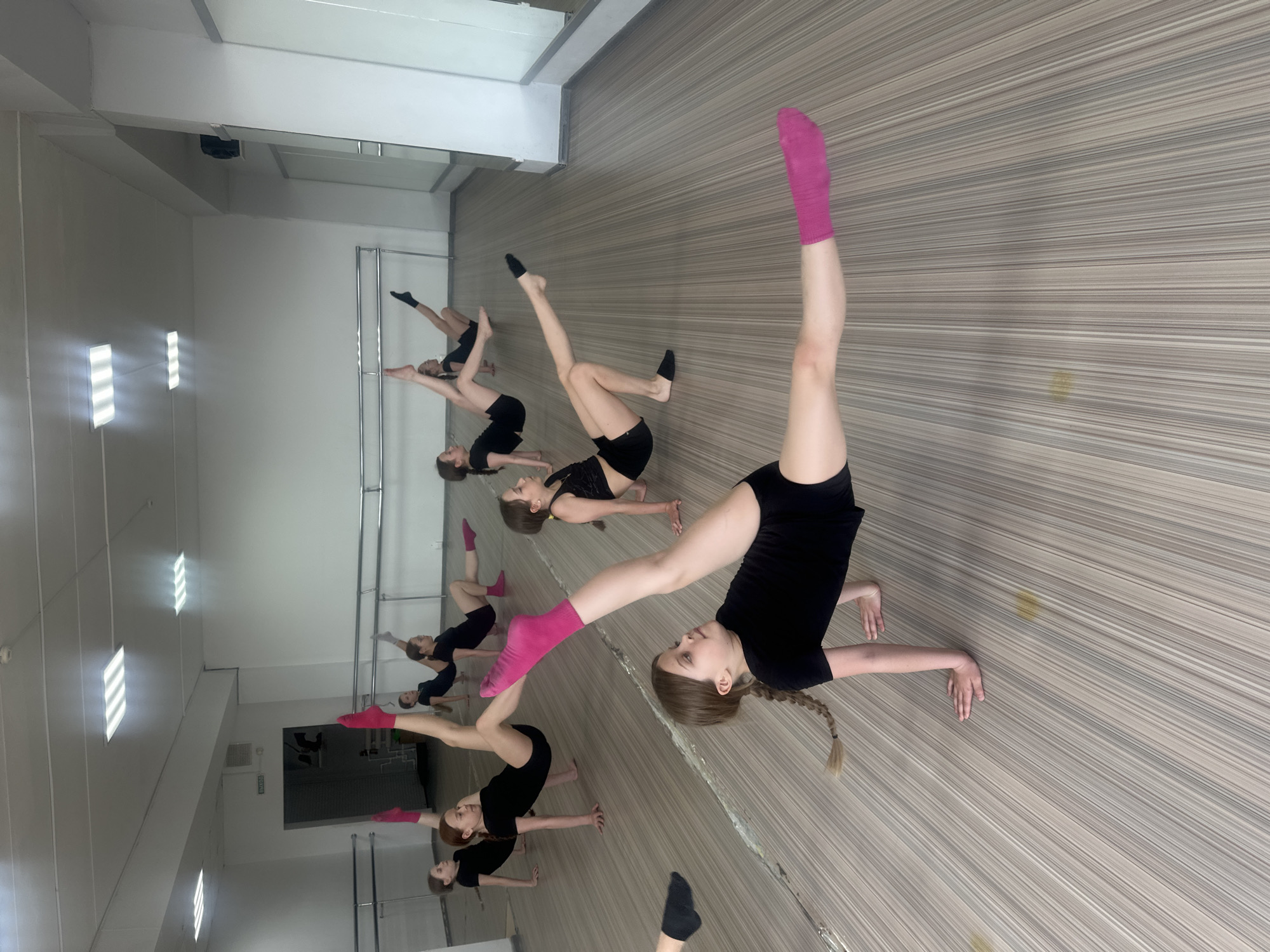 A dance class is taking place in what appears to be a studio. Seven young women are visible, dressed in black leotards and some wearing socks, one with pink socks, engaged in a flexibility exercise on the floor. They are mirrored in a reflective surface that suggests a dance studio setting with barre work reflected in the mirror, with additional girls in the background. The floor has a striped pattern. The room appears to be well-lit, with modern lighting fixtures on the ceiling. The location is unknown.