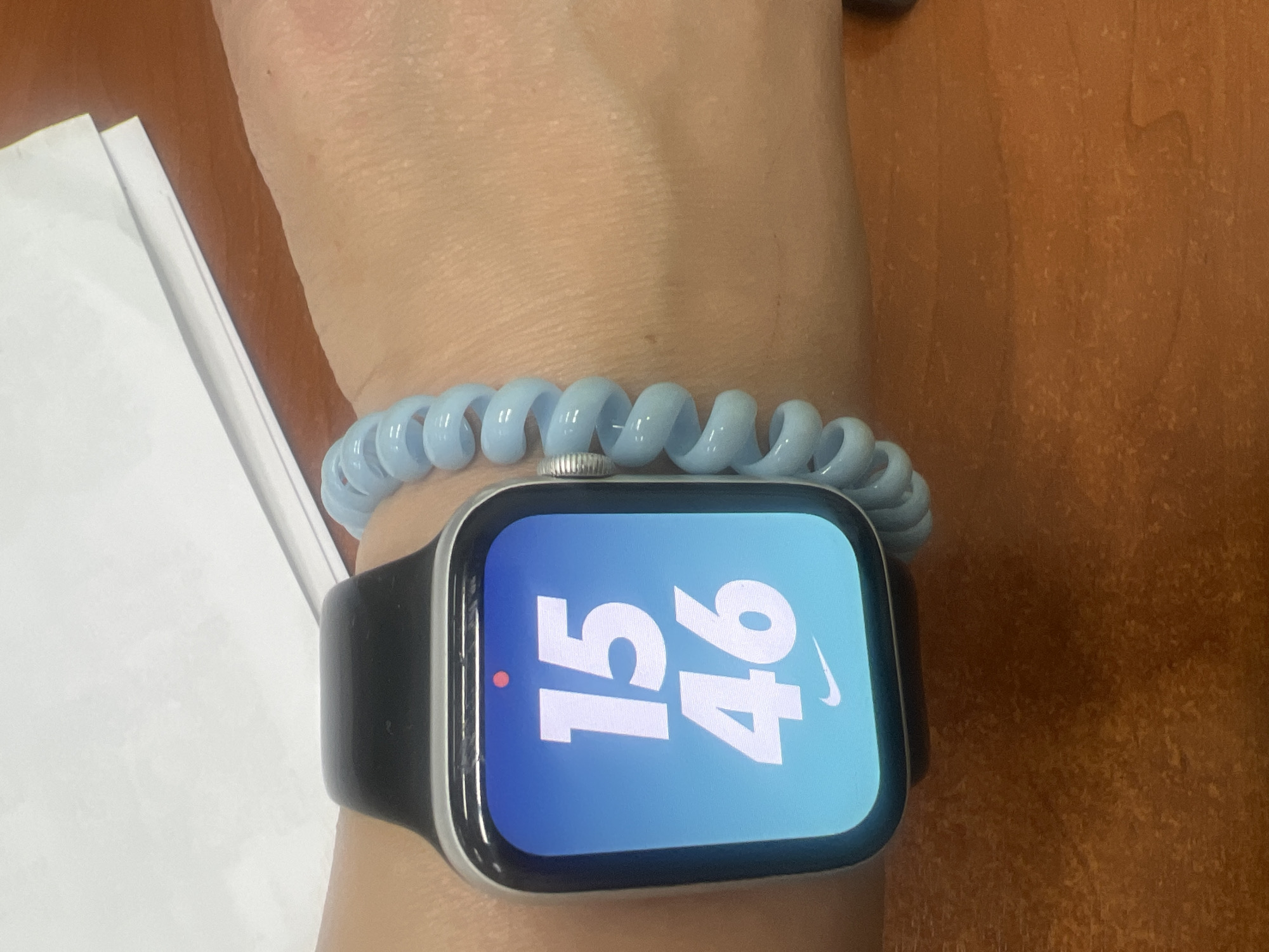 The image shows a close-up of a person's wrist wearing a smartwatch and a light blue spiral bracelet. The smartwatch display shows the time as 15:46 and features the Nike logo. The background appears to be a wooden desk. The overall scene is likely indoors, given the lighting and desk surface. The image is a still shot, so no activity or event is taking place other than the display of time. The color of the bracelet and the bright display of the watch are notable details. There is no information about the weather or time of day beyond what is shown on the watch face. The location context of Chaikovsky, Russia is not visually evident in the image.