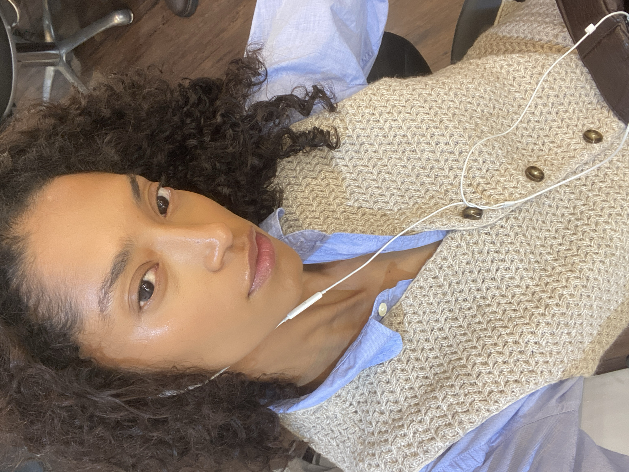 A woman with curly dark hair and light brown skin is facing the camera. She's wearing a light blue button-down shirt and a beige knitted vest with three brown buttons. White earbud headphones are connected to her ears. The background shows part of a chair and wooden flooring. This image does not contain information indicating it was taken in São Paulo, Brazil.