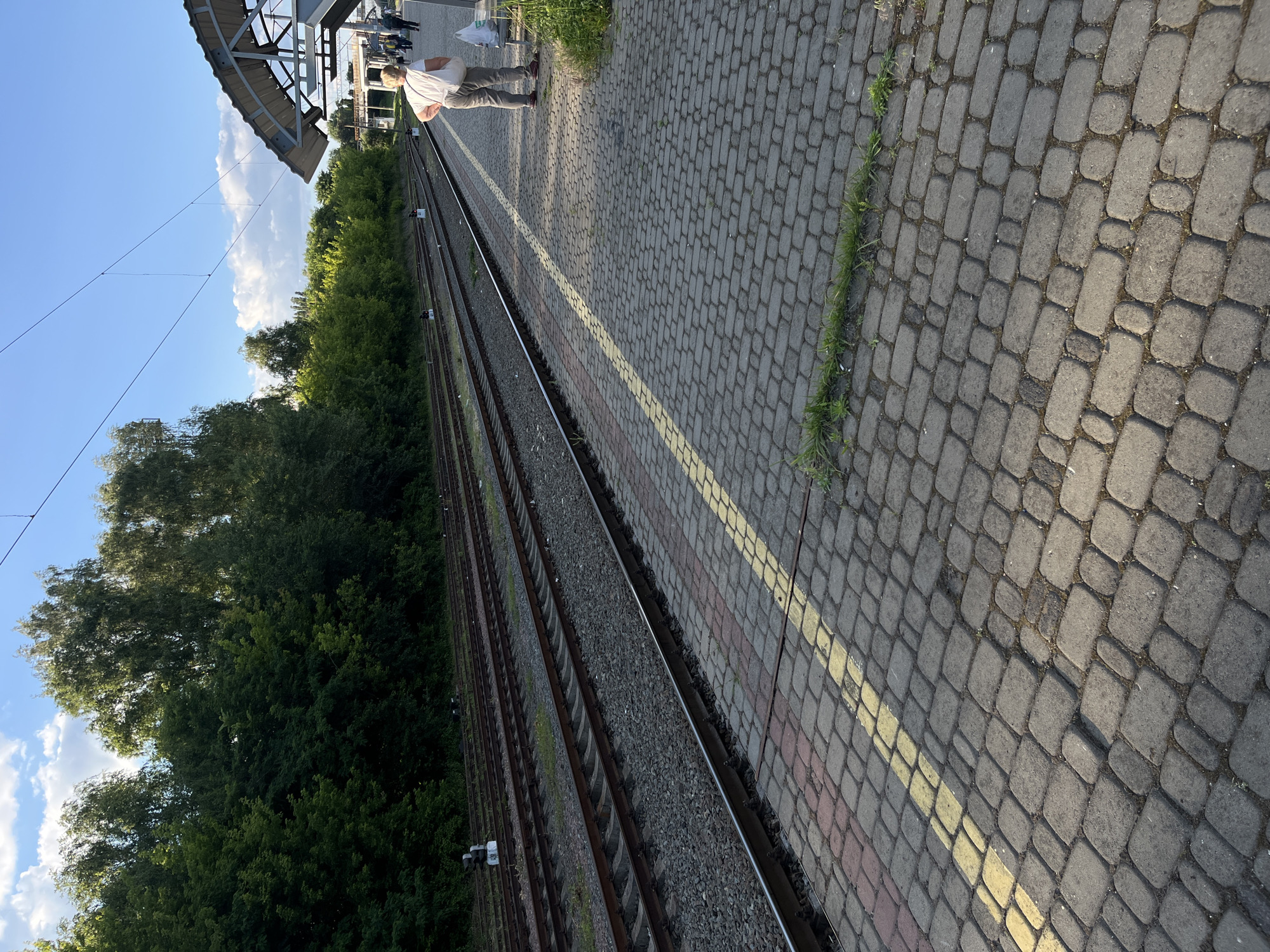 The image depicts a train station platform in Kyiv, Ukraine, with multiple train tracks visible. In the foreground, a person wearing light-colored pants and a white t-shirt stands with their back to the camera, holding a white bag. They appear to be waiting on the platform, which is paved with cobblestones and marked with a yellow safety line. The platform is adjacent to a row of train tracks, beyond which is a dense line of trees. In the background, a train station shelter with a curved roof is visible, and more people can be seen walking on the platform. The sky is blue with scattered clouds.