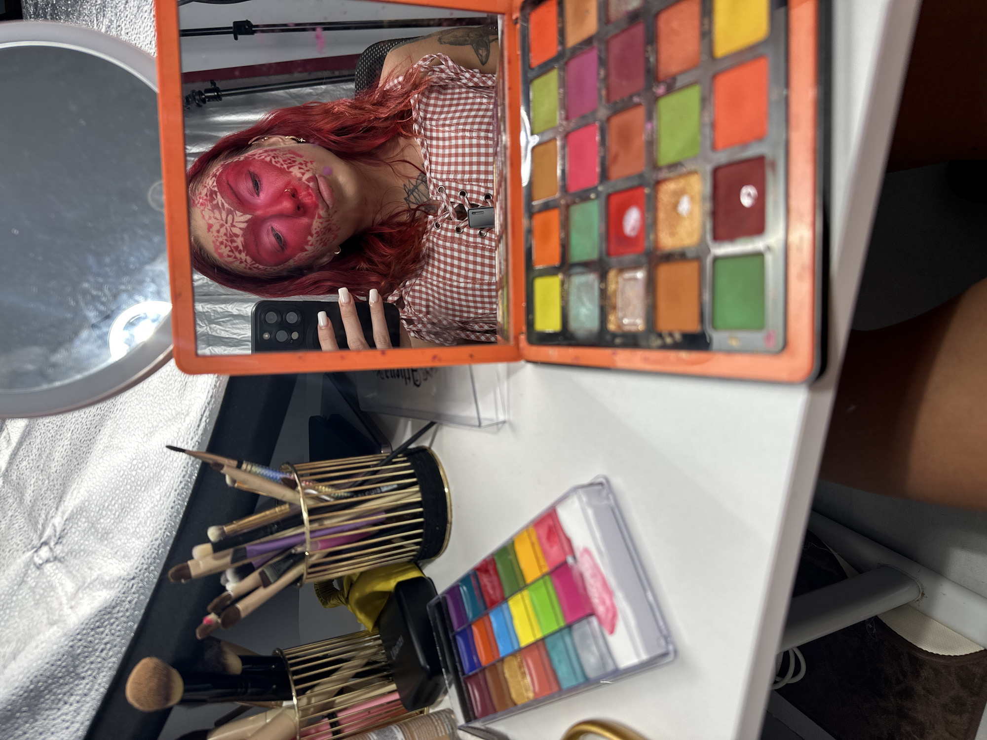 The image shows a person with vibrant red hair, wearing a red and white checkered top with a lace-up detail. They have applied dramatic pink makeup to their face, with intricate patterns drawn around their eyes and forehead. Their eyes are closed, and they are holding a smartphone.
In the foreground, a white table holds various makeup items. To the right of the person's reflection, there is an open makeup palette filled with numerous colorful eyeshadows, ranging from bright pinks and oranges to greens and blues. Below this, a rectangular palette of colorful face paints is visible. To the left, several makeup brushes are stored in a gold cylindrical holder.
The scene appears to be a personal makeup setup, possibly for a photoshoot or performance, given the elaborate face paint and the array of professional-grade makeup. The lighting suggests an indoor environment, possibly a home studio or a dressing room. The overall impression is one of creativity and artistic expression. The location cue "North Kuta, Indonesia" is not visually represented in the image.