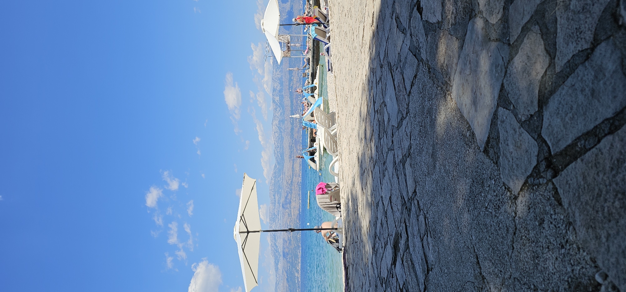 In the city of Supetar, Croatia, a paved stone walkway with visible cracks and gravel transitions to a sandy beach lined with lounge chairs and umbrellas. The turquoise waters of the Adriatic Sea stretch towards a distant, hazy mountain range under a clear blue sky dotted with scattered white clouds. Several white lounge chairs and a large white umbrella are positioned on the beach, some occupied by unseen individuals. A person in a red swimsuit stands near the water's edge, facing away from the camera. In the distance, a pier extends into the sea, with more lounge chairs and a small building. Several pedal boats are moored near the pier. The overall scene depicts a serene beach resort environment on a sunny day.