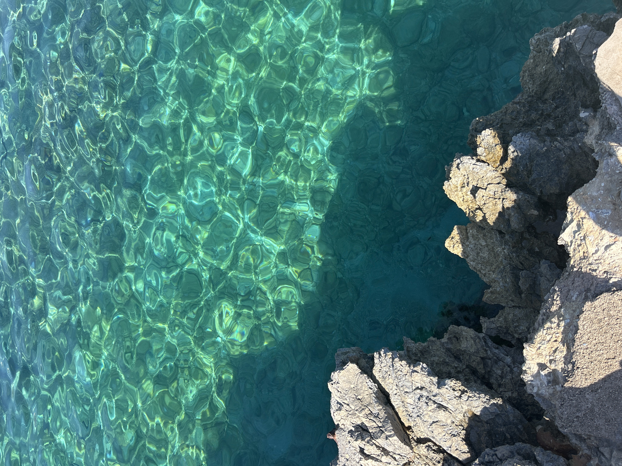 The image shows a close-up, overhead view of clear, turquoise water with light reflecting off the rippling surface, creating a mosaic of patterns. To the left and bottom of the frame, rough, grey and tan rocks jut out into the water, casting shadows. The scene appears to be a rocky coastline. The location is identified as Cala Lliteres, Spain. There are no people, animals, or man-made objects visible in the image.