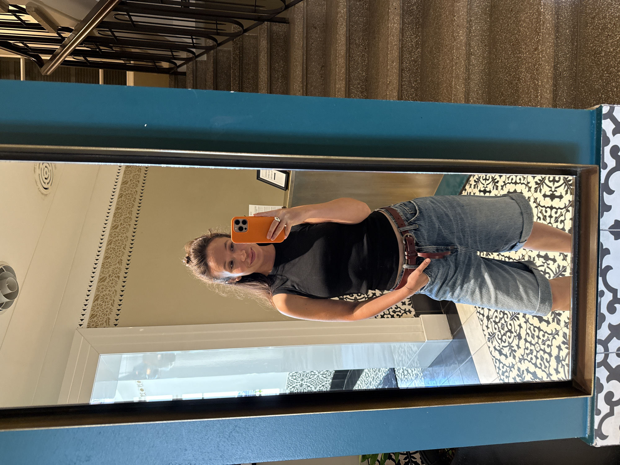 A woman takes a mirror selfie indoors, holding an orange smartphone in her right hand. She is wearing a black sleeveless top and denim shorts. The background shows a patterned tiled floor and a staircase. The mirror is framed in blue and gold. The photo appears to have been taken in Tel-Aviv, Israel.