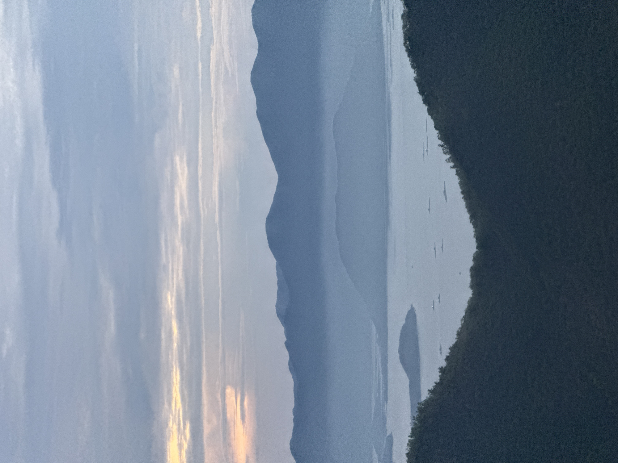 The image depicts a hazy landscape in Bac Nha Trang Ward, Vietnam. There is a lake in the center, flanked by mountains. The sky is cloudy, with a hint of sunrise or sunset on the left. The mountain on the right is covered in dense vegetation.