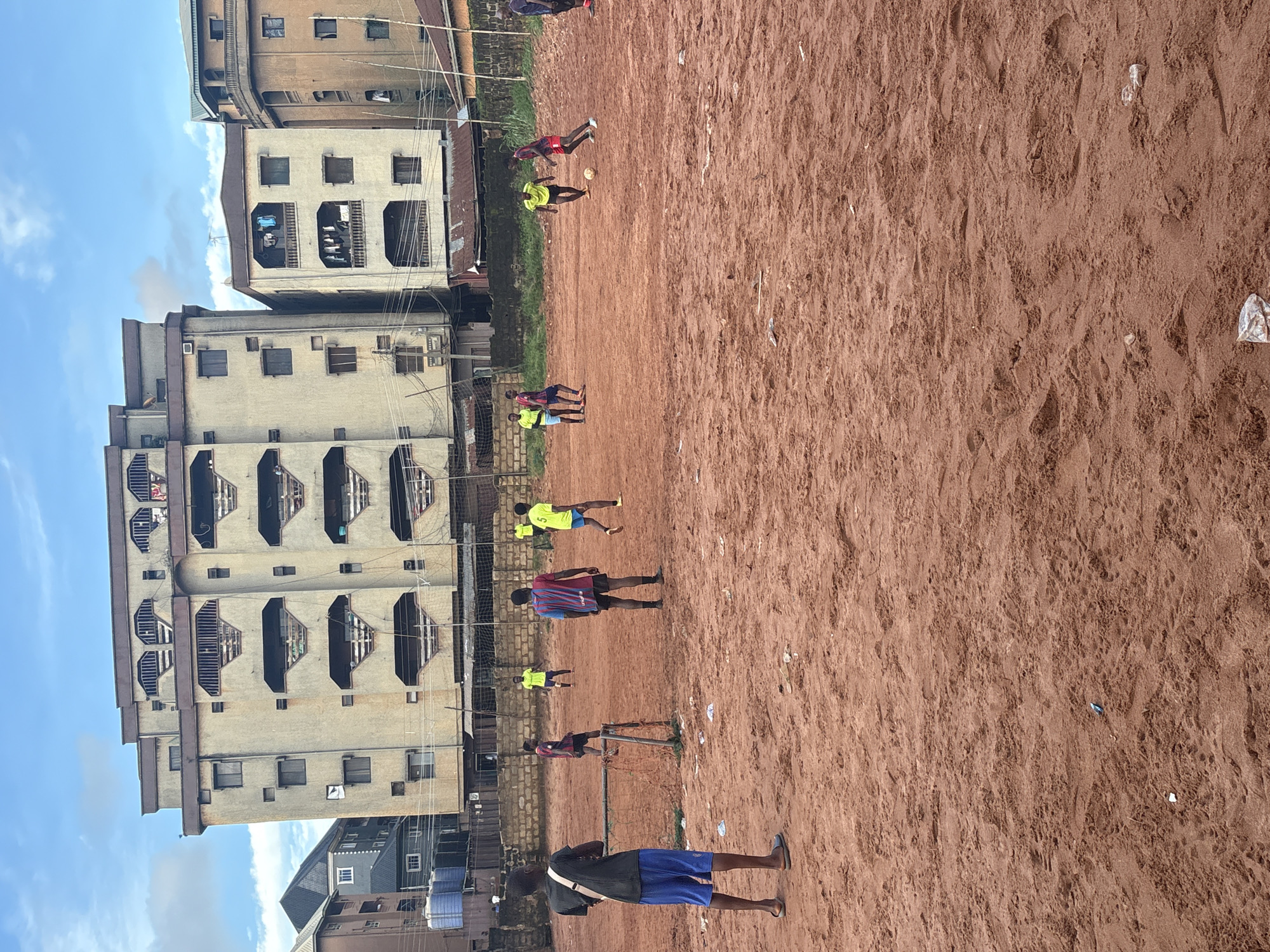This image depicts an outdoor scene on a dirt field, likely a makeshift soccer pitch, in Onitsha, Nigeria. The ground is covered in reddish-brown dirt, with scattered debris including plastic bottles and wrappers. In the background, several multi-story buildings stand, some appearing residential with laundry hanging from balconies. The sky is partly cloudy.
Several young men are engaged in a game of soccer. One person in the foreground, wearing a dark t-shirt and blue shorts, stands facing away from the camera, seemingly observing the game. Other players are spread across the field, wearing various colored jerseys and shorts. Two individuals in bright yellow shirts and blue shorts are visible, one near the center and another further back. Another player in a red and blue striped jersey is near the center of the field. Two figures in red shorts and yellow/green tops are interacting with a soccer ball in the mid-ground, one kicking it. A goalpost with a net is visible on the left side of the image. The overall impression is a casual game being played in an urban environment.