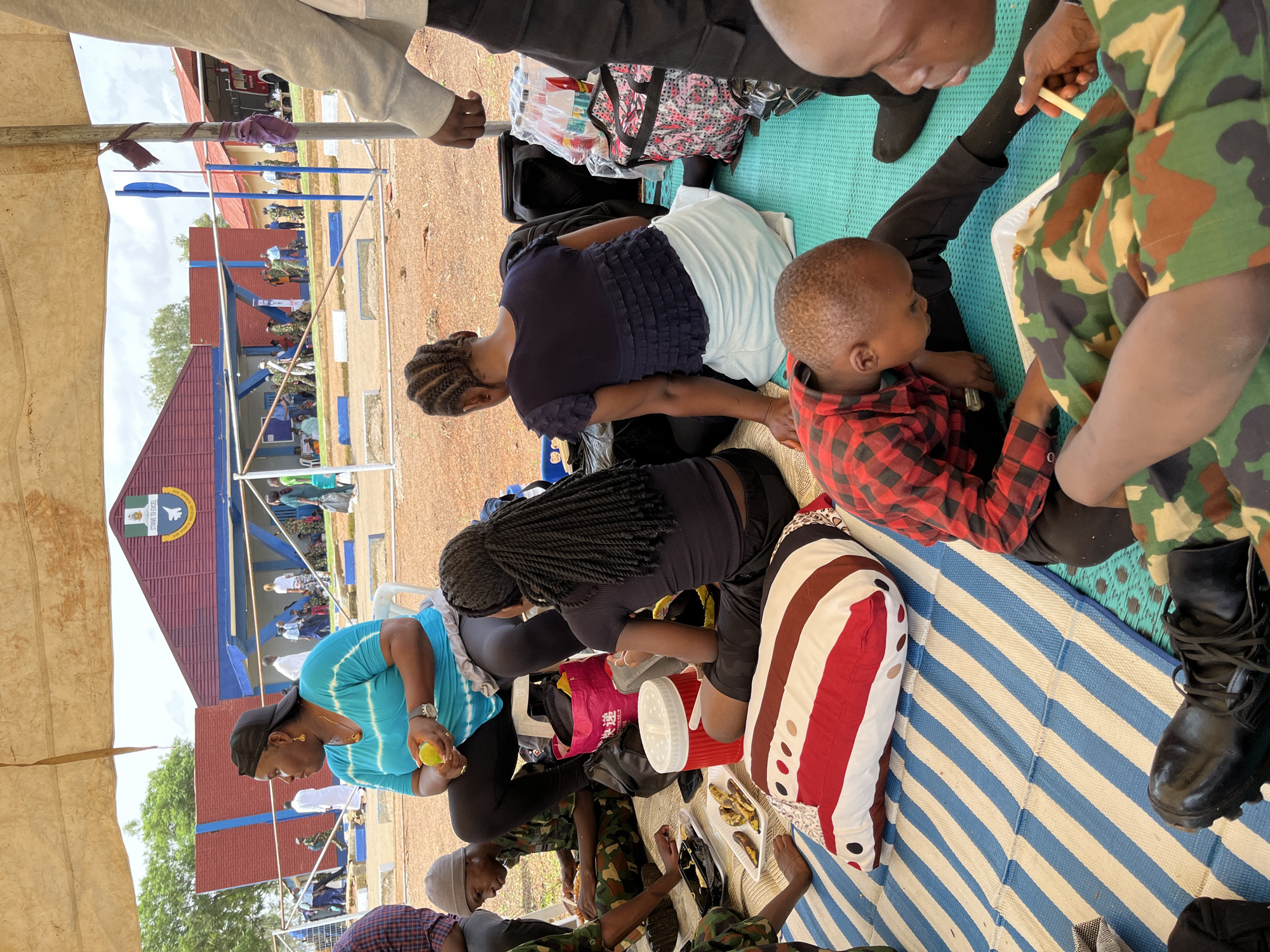 In Bar Jirgi Summa, Nigeria, a group of people are gathered outdoors under a temporary shelter. The ground is covered with blue and white striped mats and a teal mat, with a striped pillow and various bags scattered around. In the foreground, a young boy in a red and black plaid shirt is eating from a white plastic container. To his right, an adult in camouflage clothing is holding a utensil and appears to be eating. In the middle ground, a woman in a blue tie-dye shirt is seated, holding a yellow fruit. Next to her, a woman with braided hair is seated facing away from the camera. Behind them, several people in camouflage uniforms and other attire are visible, walking or standing in front of a building with a red roof and blue trim. The building has a large emblem on its facade and what appears to be a fire truck visible in an open bay on the right side of the frame. The overall scene suggests a gathering or event taking place in an outdoor, possibly institutional setting.