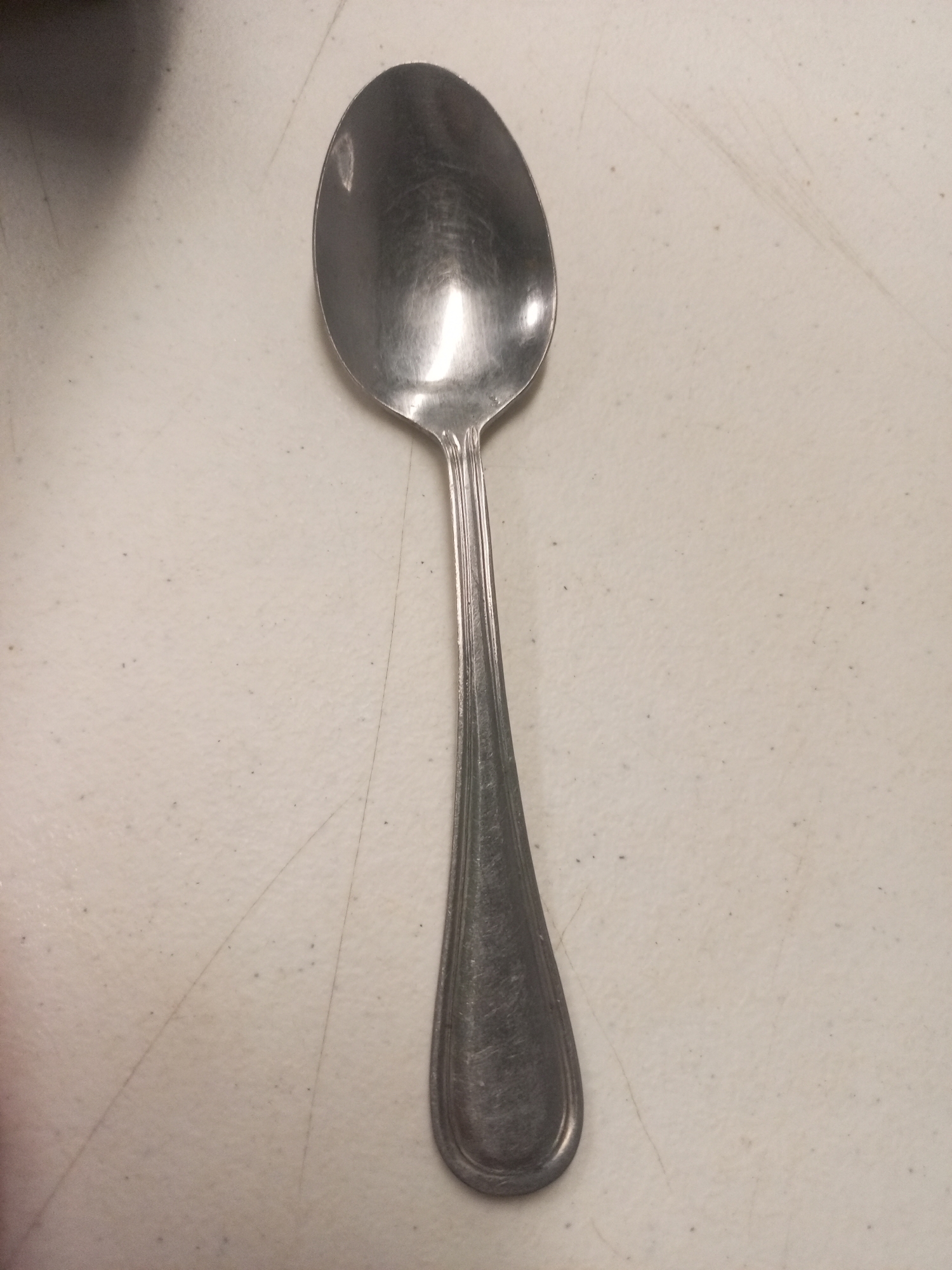 A silver spoon lies on a textured, light-colored surface. The spoon is oriented vertically in the image, with the bowl at the top and the handle extending down. The bowl is oval-shaped and reflects light. The handle tapers from the bowl, widening slightly before ending in a rounded shape. No other objects or people are visible. There is no discernible information to place this image in Juan Griego, Venezuela.