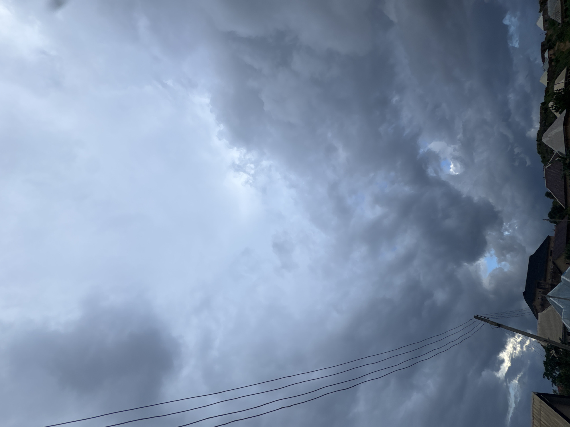 The image captures a dramatic sky filled with dark, heavy storm clouds over a residential area. In the foreground, power lines stretch diagonally across the frame, supported by a wooden pole. Below the sky, several houses with pitched roofs are visible, their structures silhouetted against the ominous clouds. The overall atmosphere is one of impending weather change, with the sky dominating the scene.