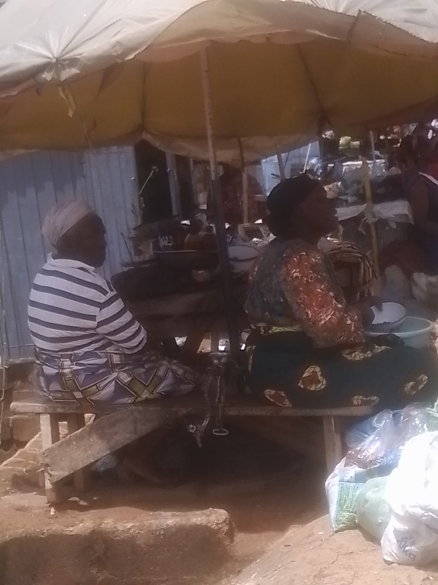 This image depicts a market scene in Gwong, Nigeria, likely during the daytime given the bright lighting filtering through a large, yellow market umbrella that covers the setup. Two women are seated on a wooden bench under the umbrella, facing away from the camera. The woman on the left wears a light-colored head wrap and a horizontally striped t-shirt in white and blue. She also wears a patterned skirt in blue, white, and yellow. The woman on the right has dark hair and is wearing a patterned, long-sleeved blouse with floral motifs in red, white, and green, over a dark green skirt with a yellow and white patterned border and heart-shaped motifs.
In front of the women, there is a table or surface where various objects are placed, including bowls and possibly food items or goods for sale. One bowl in front of the woman on the right appears to contain a white substance. To the right of the bench, several large bags are stacked, some translucent and containing what looks like dried goods or produce, and others opaque, one appearing light green and another white. The background includes a corrugated metal structure, suggesting a permanent or semi-permanent stall. The ground appears to be made of dirt and possibly some rough-hewn stones or steps in the foreground. The overall atmosphere seems to be one of everyday commerce.