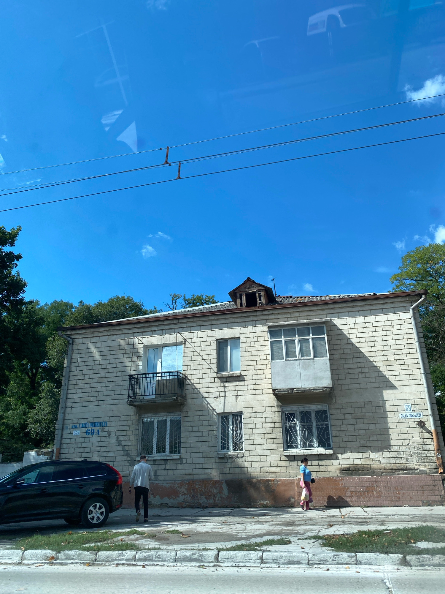 The image captures a street view in Chișinău, Moldova, focusing on a two-story brick building with a light-colored facade. The building appears to be an apartment complex, with multiple windows and a small balcony. Signs on the building indicate street names and numbers; one sign reads "STR. CALEA ORHEIULUI 69 / 69A". A black SUV is parked on the left side of the street, and two individuals are walking on the sidewalk in front of the building. The sky is bright blue with some scattered clouds, and overhead power lines are visible. The overall scene suggests a typical urban environment on a sunny day.