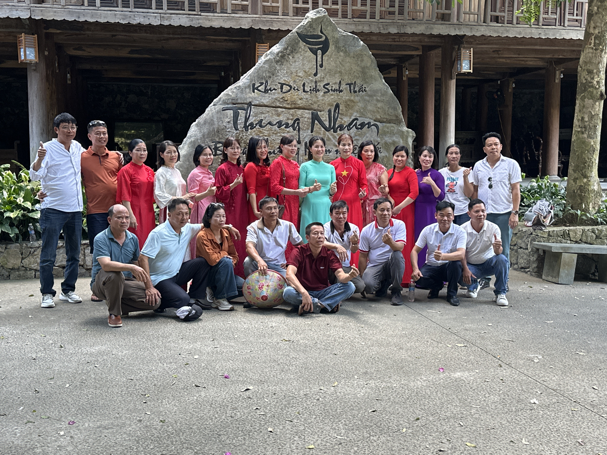 A group of approximately twenty-five people, mostly adults, are gathered outdoors in front of a large rock inscribed with Vietnamese text and a stylized bird logo. The rock appears to be a landmark at the "Khu Du Lich Sinh Thai Thung Nham" tourist site. The individuals are dressed in a mix of casual and traditional attire, with many women wearing red or pink outfits and several men in blue shirts and jeans. Some are standing, while others are crouched or seated on the ground. The setting suggests a communal gathering or tourist activity within a lush, natural environment, possibly a park or resort, indicated by the presence of trees and stone seating in the background. The lighting suggests it is daytime, and the overall atmosphere is relaxed and social. The architecture visible in the background, with its wooden beams and balconies, hints at a traditional Vietnamese building style.
