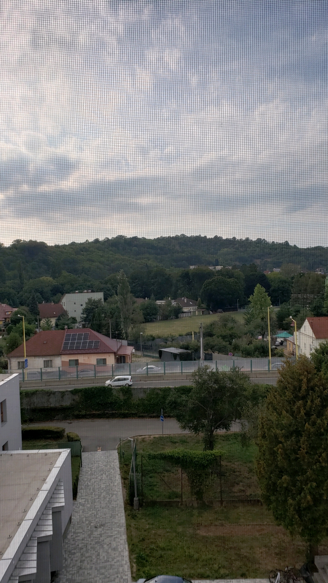 The image depicts a view of a residential area with a hill covered in trees in the background. In the foreground, there is a street with a white car driving on it, separated from a residential building by a green fence. A house with solar panels on its roof is visible on the left side of the street. The sky is overcast. The scene appears to be a typical suburban or semi-urban environment.