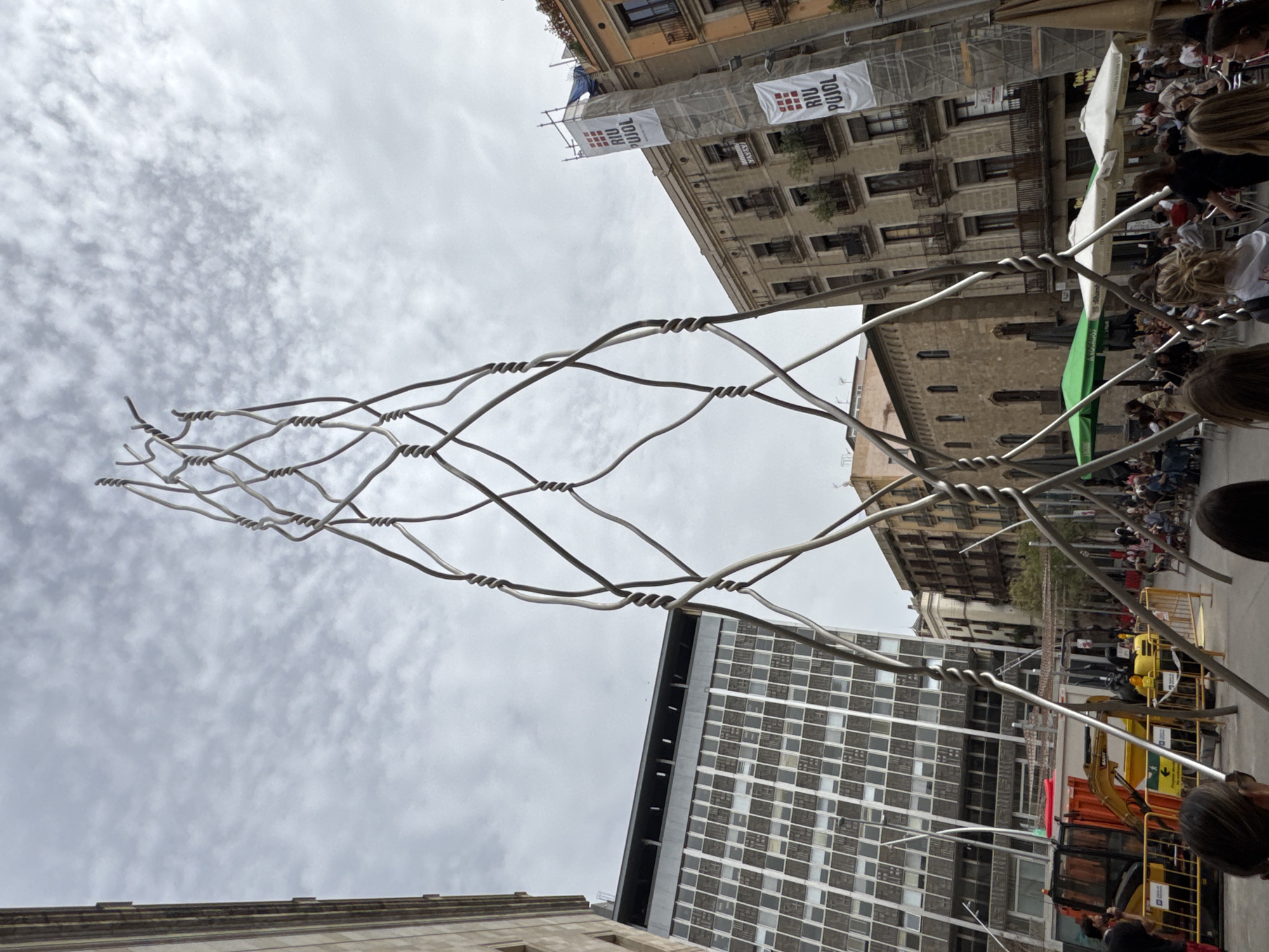 A low-angle, medium shot depicts a street in Barcelona, Spain. An art installation comprising intertwined, twisted metal wires dominates the foreground, stretching upwards against a cloudy sky. Below the installation, a large crowd has gathered, lining the street and partially obscured by the structures of the installation.
Buildings of varying architectural styles are visible on both sides of the street. The building to the left is modern and high-rise with a grid-like pattern of windows. Across the street, the architecture is older, with balconies and hanging banners reading "RIU PUJOL" partially obscured by scaffolding.
There's street equipment and machinery visible on the right side of the street, partially obstructed by people. The day appears overcast.