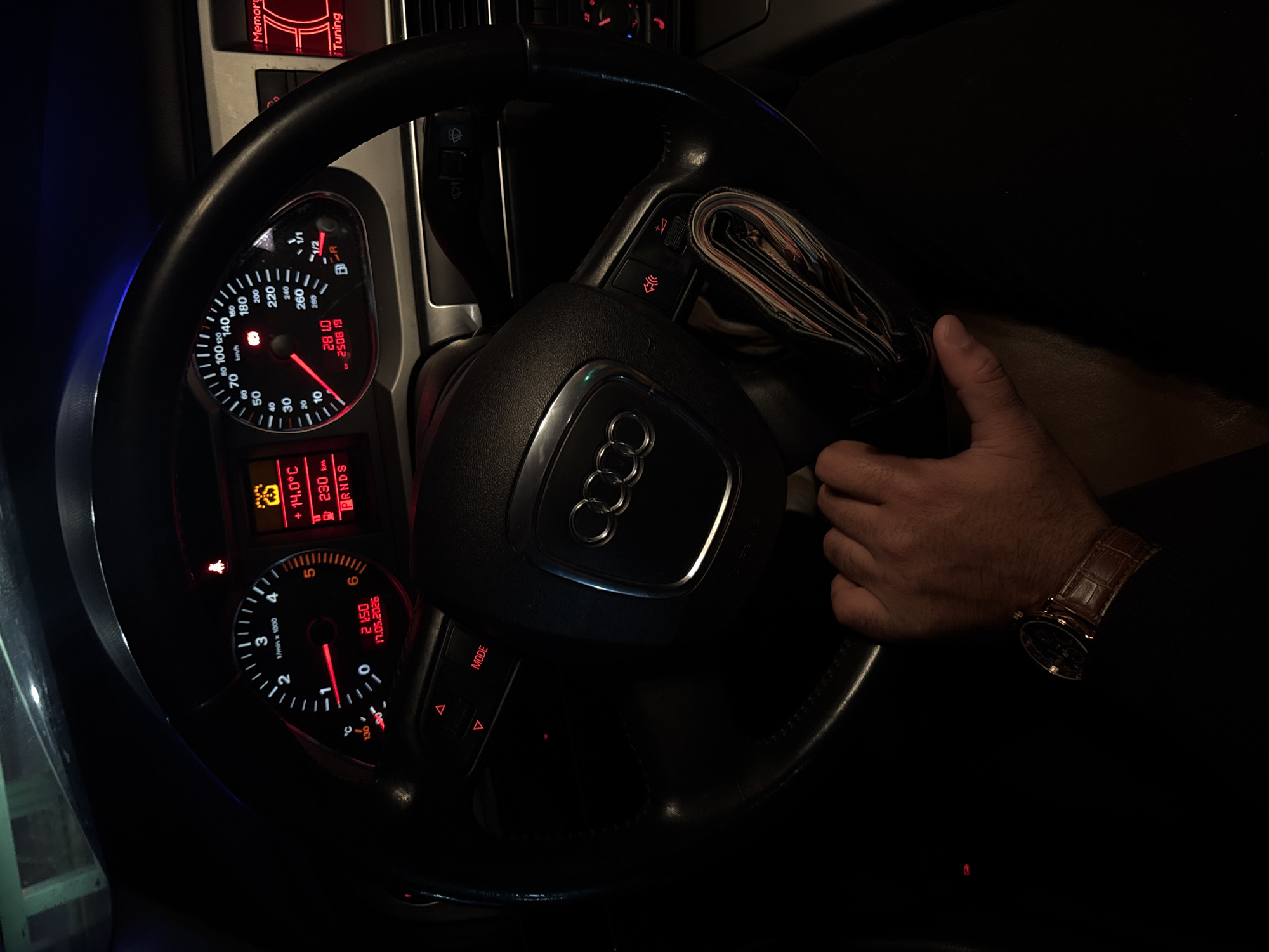 A dimly lit car interior is visible in the image. An Audi steering wheel dominates the scene, with the Audi logo prominently displayed in the center. The car's dashboard instruments are illuminated in red, displaying information like speed, RPM, time, date, and temperature. The time displayed is 21:50 on 17.05.2026. A man's hand wearing a brown leather watch is visible on the steering wheel, suggesting the driver's presence. A wallet, likely containing money, is positioned next to the steering wheel. The setting implies the car is stationary or being driven slowly in low light conditions. The location is not discernible from the image.