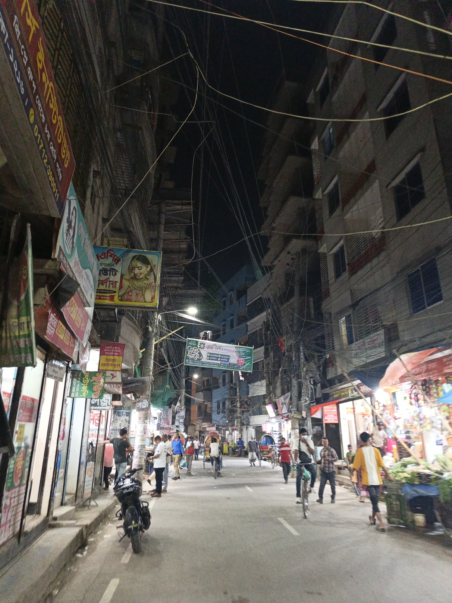 This is a street scene in Dhaka, Bangladesh. The narrow street is lined with buildings, many plastered with signs and advertisements in Bengali. Power lines crisscross overhead in a chaotic web. Pedestrians walk along the street and on the sidewalks. Some are on bicycles and rickshaws. A parked motorcycle is visible near the left edge of the frame. Shops are open, with one featuring a vegetable stand. The sky is dark, suggesting it's nighttime, with streetlights illuminating the scene. The image portrays a typical, bustling urban environment in Dhaka.