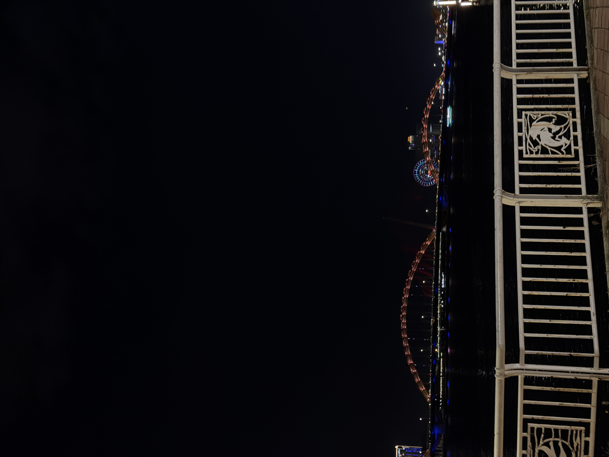 The image captures a nighttime scene in Da Nang, Vietnam. The composition is dominated by a large, dark body of water, reflecting the vibrant lights of a cityscape and an illuminated Ferris wheel in the distance. On the right side of the frame, a white metal railing with horizontal bars and decorative panels featuring stylized animal figures runs vertically. The railing appears to be part of a promenade or viewing area along the waterfront.
The city lights are a mix of warm white and colorful hues, outlining buildings and bridges, creating a festive and lively atmosphere. The Ferris wheel, a prominent feature, is outlined with lights, suggesting it is operational and a popular attraction. The water's surface is still, providing a clear, albeit slightly distorted, reflection of the illuminated structures.
The time of day is clearly night, indicated by the darkness of the sky and the reliance on artificial lights for illumination. There are no discernible weather conditions, but the reflections suggest calm water. The presence of the Ferris wheel and the waterfront development point towards a tourist or entertainment district. The overall mood is one of urban vibrancy and nocturnal beauty. No visible text is present in the image.