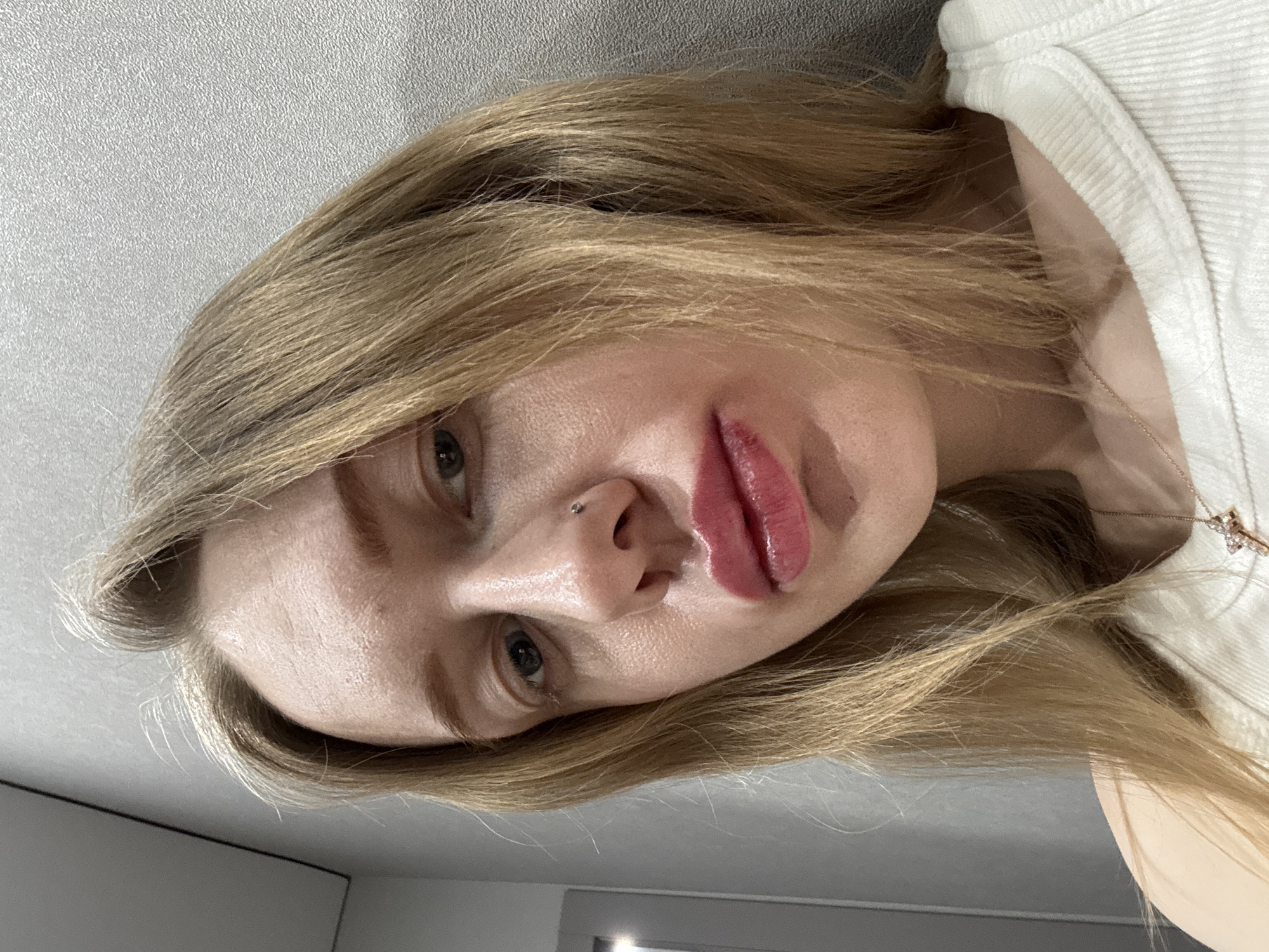 The image presents a close-up portrait of a light-skinned adult female in an indoor setting in Rostov-on-Don, Russia. Her blonde, shoulder-length hair frames her face. She possesses dark-colored eyes and gazes directly into the camera. A small, silver-colored stud is visible as a piercing on the left side of her nose (subject's left). Her lips are full, coated in a pinkish-red gloss, with minor textural variations on the lower lip. Her complexion displays some redness or unevenness on the forehead and cheeks. She is wearing a light-colored, ribbed garment, likely a tank top or camisole. A delicate, gold-toned chain necklace with a small, complex pendant is visible around her neck. The background is a plain, light grey wall with a subtle texture. A dark vertical line, consistent with a door frame or room corner, is partially visible on the left, exhibiting a faint light reflection.