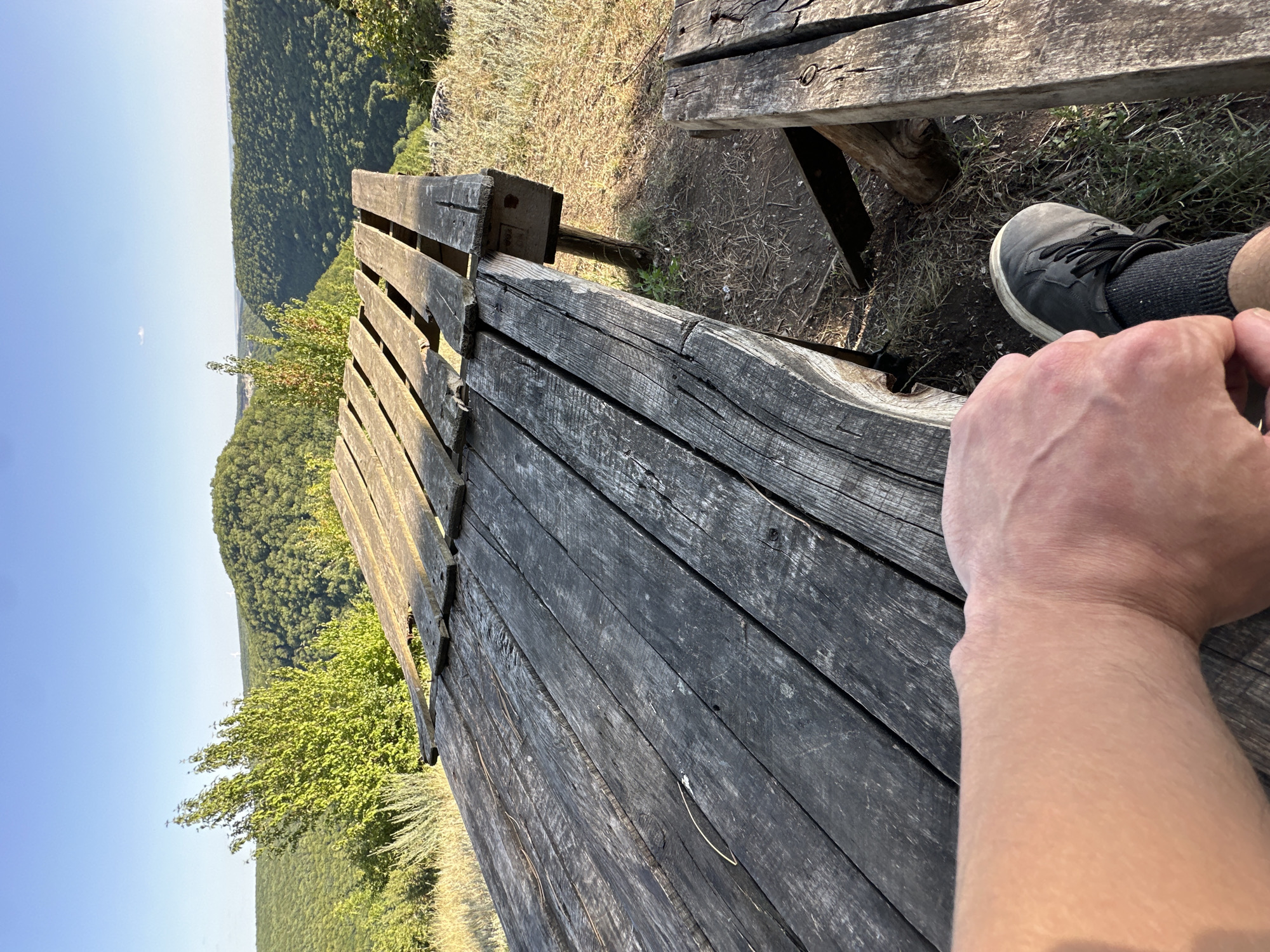 The image shows a rustic wooden picnic table in a natural setting. The table is weathered and appears to be made of rough-hewn planks. In the foreground, a person's arm and foot are visible, with the person wearing dark sneakers and socks. The background reveals a scenic landscape with rolling, tree-covered hills under a clear blue sky. The location is identified as Pryvorottia Druhe, Ukraine. The scene conveys a sense of outdoor leisure or rest amidst nature.