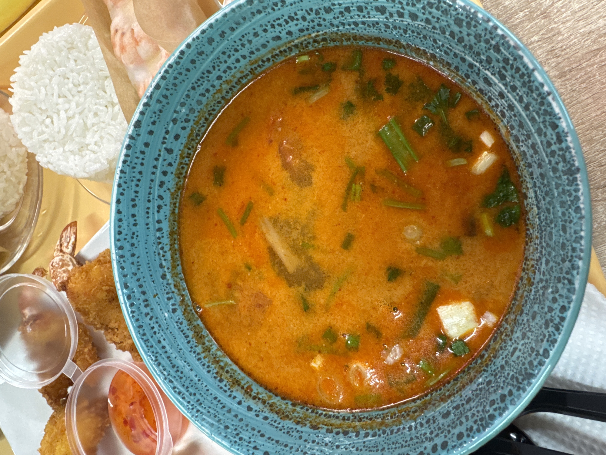 The image shows a bowl of orange soup, likely Tom Yum, with green onions and tofu in a blue bowl. There is rice and spring rolls on the side in clear plastic containers, along with a small container of orange sauce. The soup is in focus and takes up most of the frame. The image was taken from above at close range. The city and other specifics are Unknown.