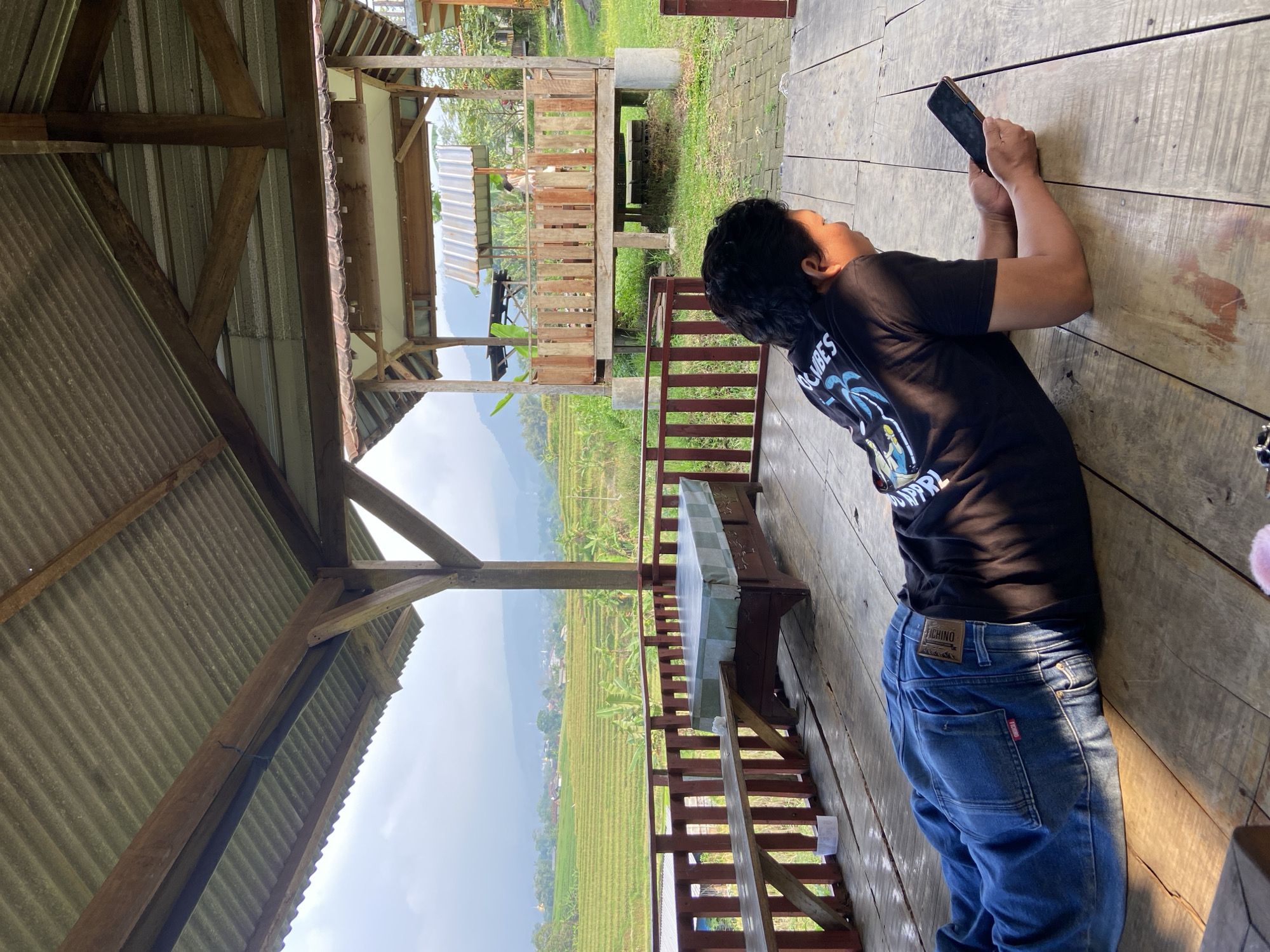 A person is lying on a wooden surface, holding a mobile phone in their hands, presumably using it. The individual is wearing a black t-shirt and blue jeans. The photo appears to be taken from an elevated angle under a sheltered, open structure with a metal roof. In the background, a rice field stretches out towards distant mountains under a partly cloudy sky. Another, smaller wooden structure is visible a short distance away. The scene is possibly located in Tamiajeng, Indonesia, as indicated in the instructions. The wooden surface the person is lying on looks like part of a platform or deck attached to the sheltered structure.