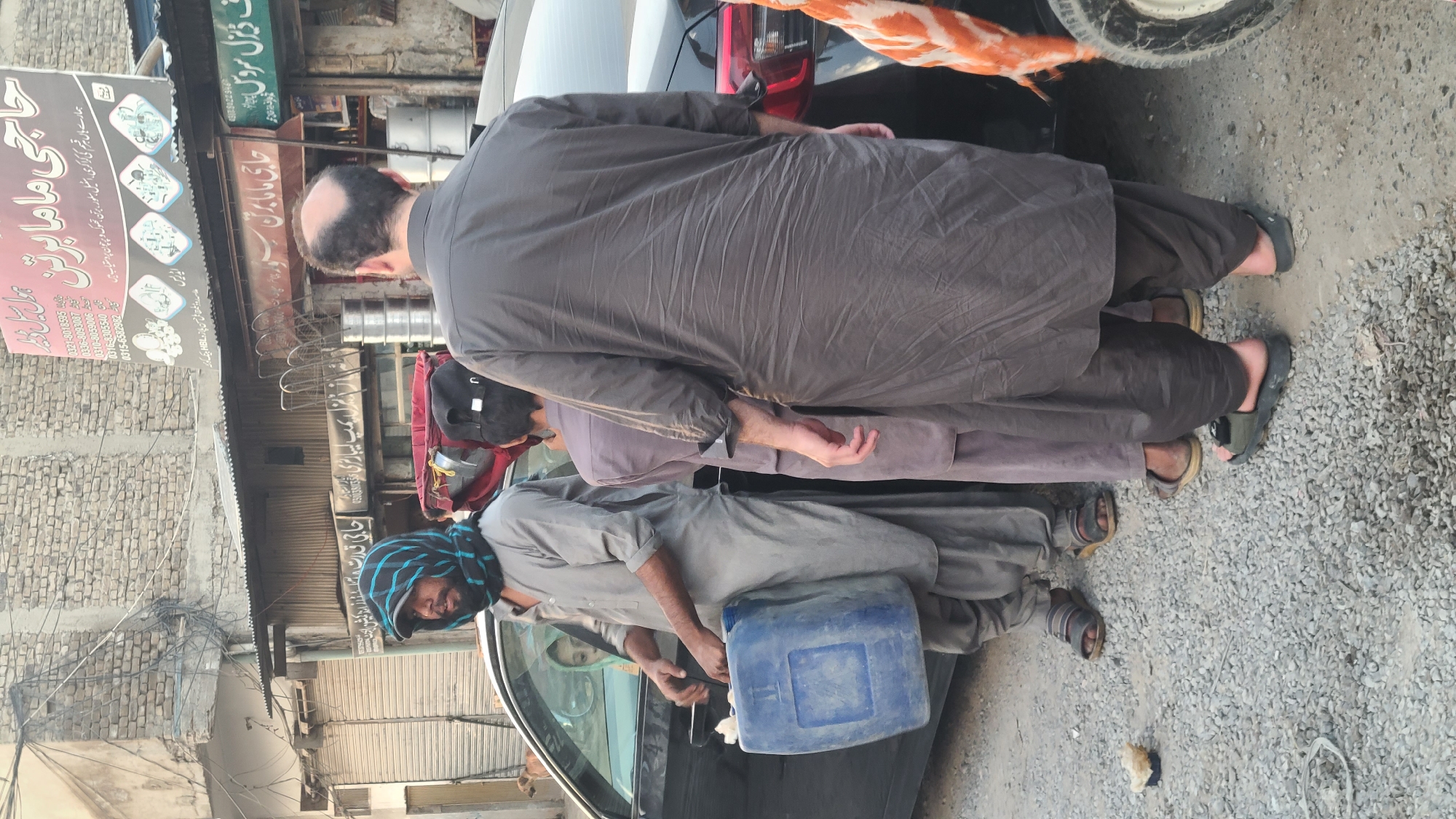 The image shows three men standing on a gravelly ground in Quetta City Tehsil, Pakistan. The man in front is wearing a light grey outfit and a blue head covering, and he is holding a blue container near a black car. The second man is wearing a darker grey outfit and a black cap. The third man is wearing a dark grey outfit and has a receding hairline. The men are standing near a building with signage in Urdu, including advertisements for "Haji Mama Bartan," a cookware store, and "F. Diesel Service." There are utility poles and wires visible in the background.