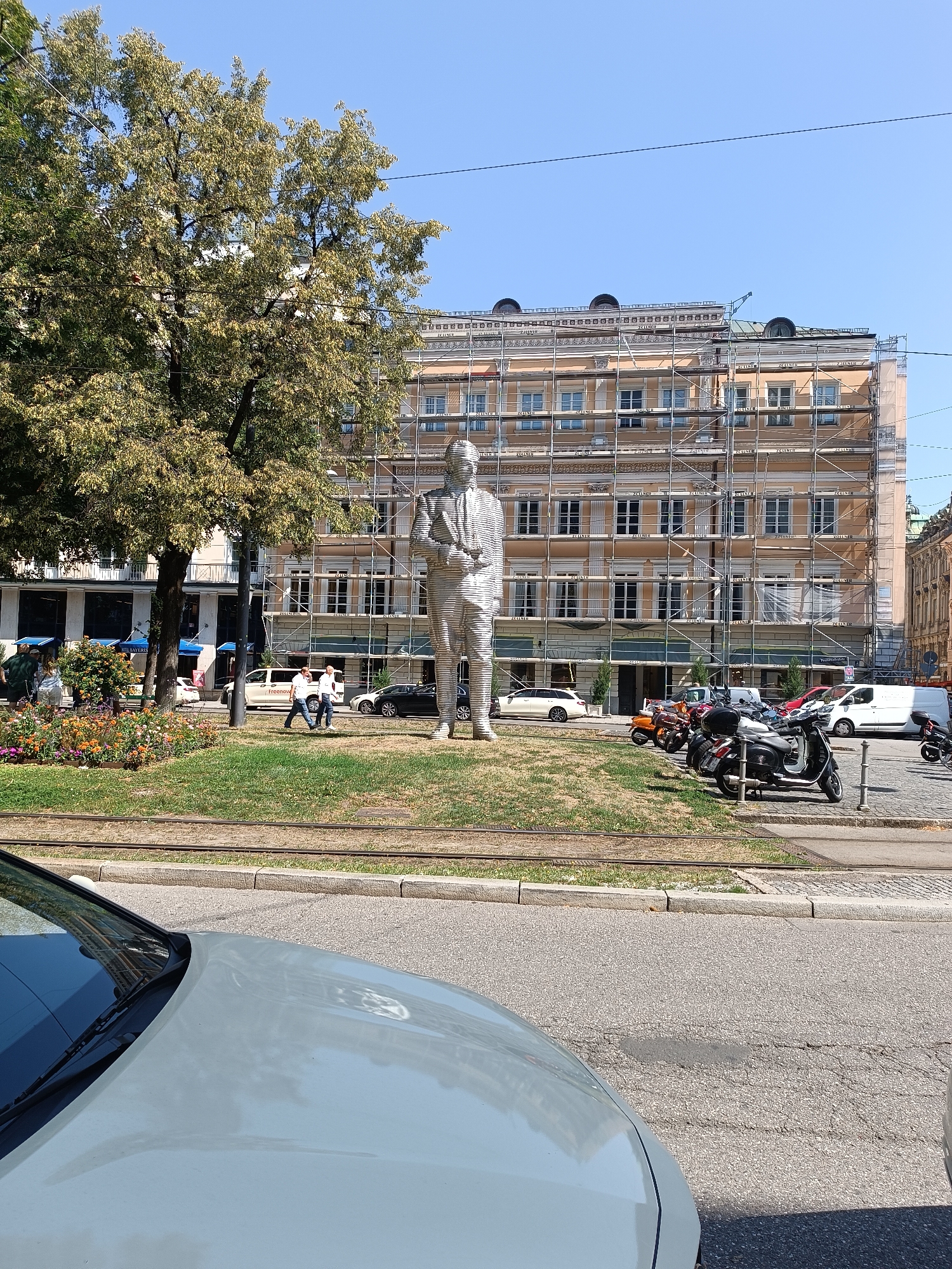 The image captures a sunny day scene in Munich, Germany, specifically at the Prinzregentenplatz. In the foreground, the hood and windshield of a light-colored car are visible. Across the street, a large, metallic sculpture of a standing man is prominent in a grassy area with flowerbeds. Behind the sculpture, a building is undergoing renovation, covered in scaffolding. Several people are walking on the sidewalk near the flowerbeds. A row of parked scooters and motorcycles is visible on the paved area. Further in the background, cars and vans are present, and tram tracks run along the edge of the grass. The sky is clear and blue.