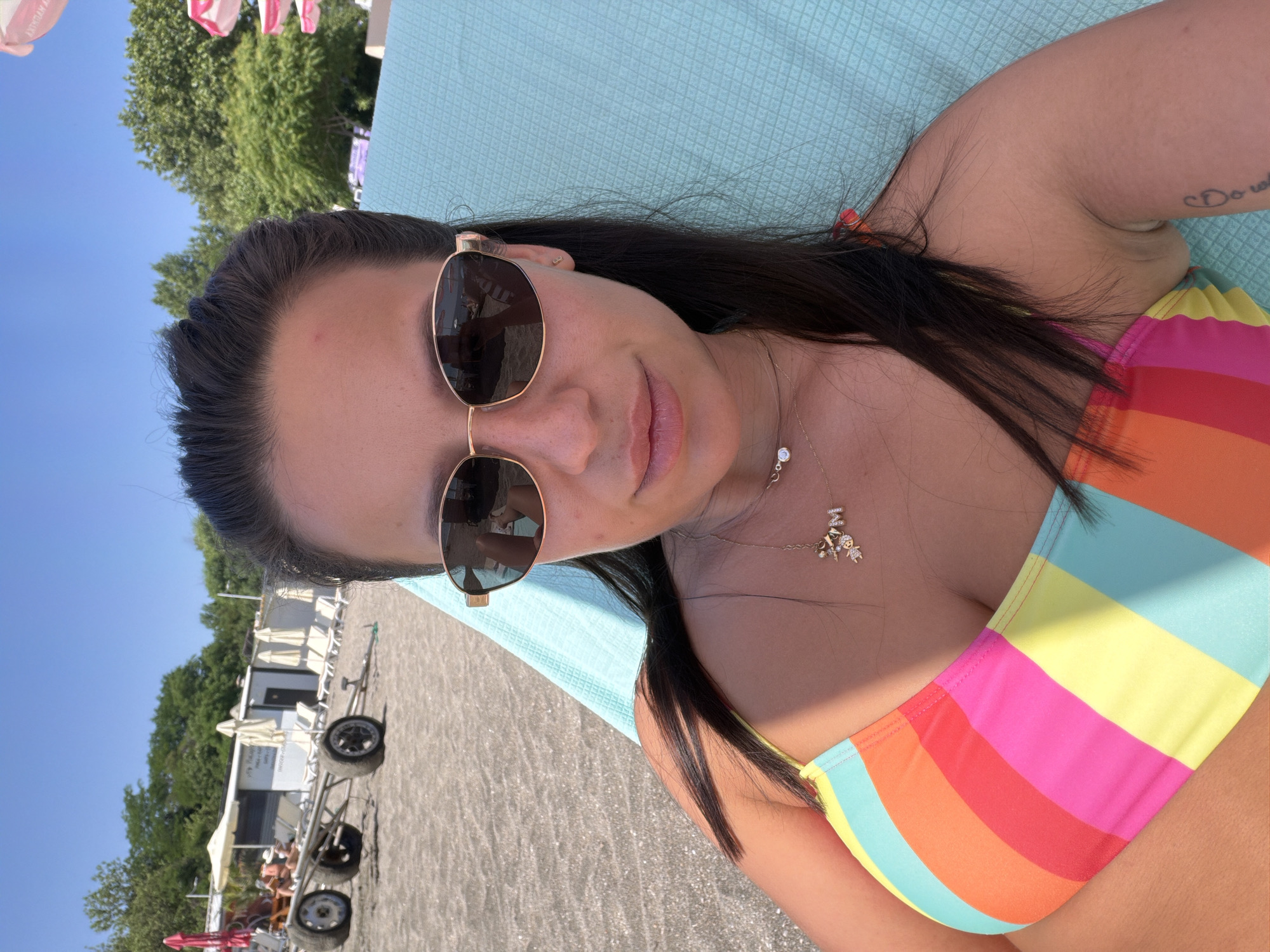 The image depicts a woman lounging on a turquoise beach chair on a sandy beach. She is wearing a colorful striped bikini and sunglasses, and has long dark hair. A gold necklace with a charm is visible around her neck. In the background, there are beach umbrellas, lounge chairs, and a white building. A cart with large wheels is also present on the beach. The sky is clear and blue, indicating a sunny day. The scene is set in Burgas, Bulgaria, specifically on the beach area.