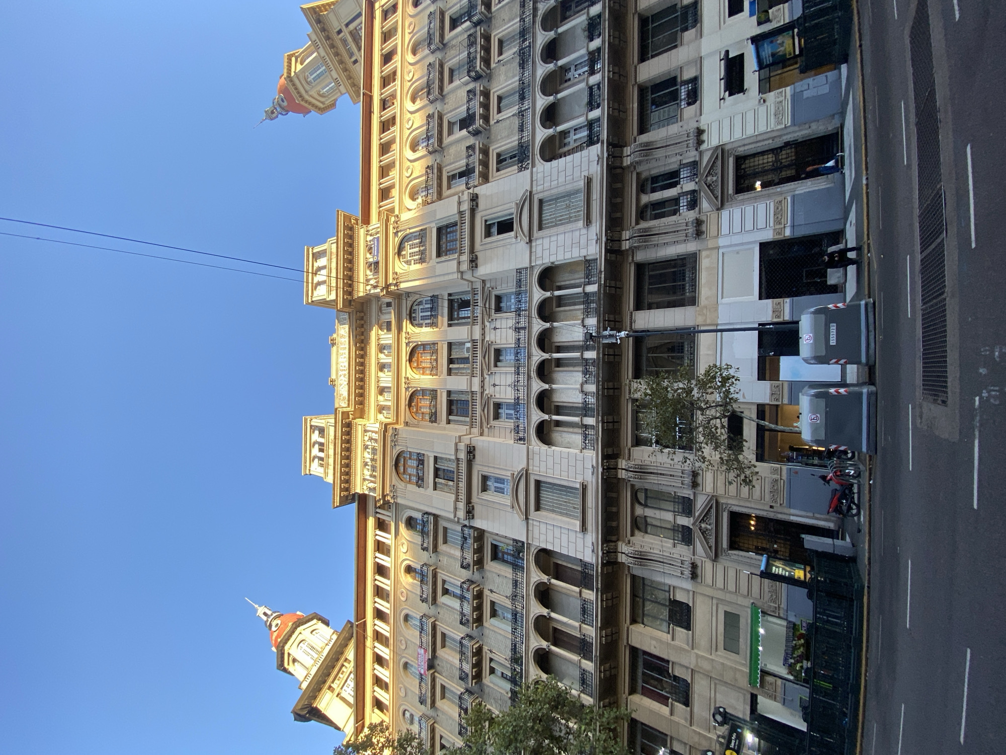 This image captures a vibrant street scene in Buenos Aires, Argentina, showcasing a grand, multi-story building under a clear blue sky. The building, a prominent subject, features a light-colored facade with intricate classical architectural details. It boasts numerous arched windows, many with ornate wrought-iron balconies and railings, and decorative cornices. Two distinctive tower-like structures with reddish, domed roofs are visible at the upper corners of the building, adding to its majestic appearance. The sunlight illuminates parts of the facade, suggesting a bright daytime setting, likely morning or afternoon.
At street level, the building houses various storefronts and entrances, including one with a dark, ornate metal gate. The pavement is made of asphalt with clear white and yellow lane markings, indicating an urban street. Notable objects on the sidewalk include a grey urban waste bin (possibly recycling) with a number "53587" and a logo, and a red bicycle parked near the building.
Several pedestrians are visible on the sidewalk; one person in a blue shirt stands near an entrance, while others are further down the street. A street pole with a sign reading "Metrobus" is visible. Overhead, thin power lines traverse the blue sky. The scene conveys the bustling yet architectural beauty of a typical day in a historic district of Buenos Aires.