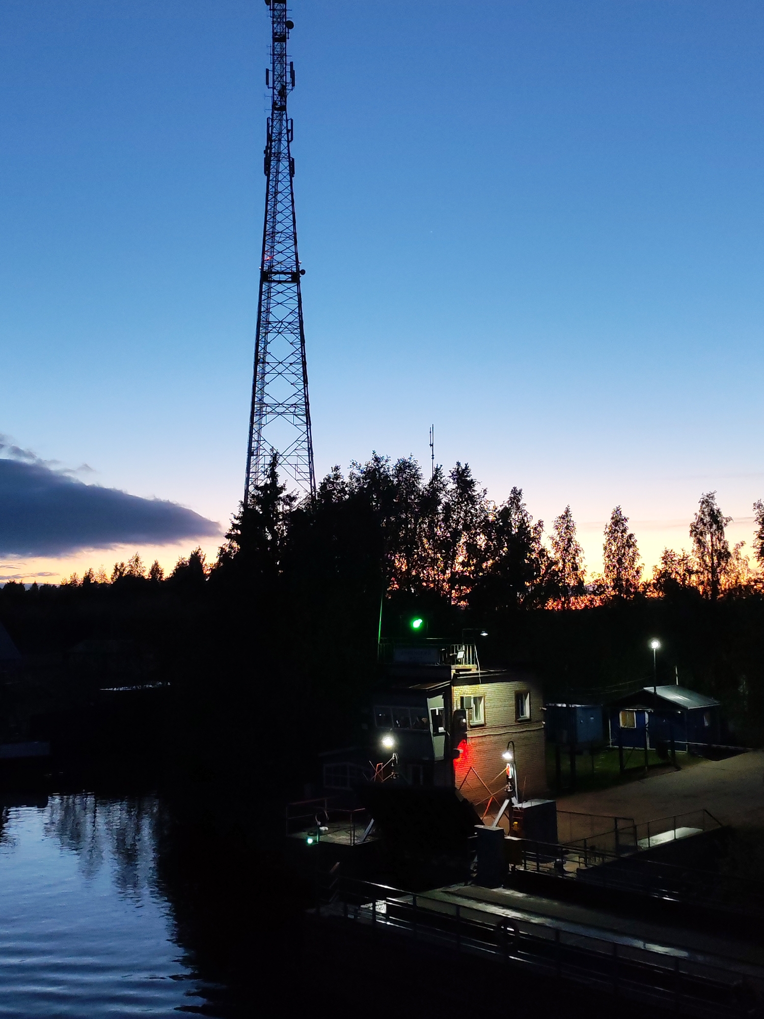 The image depicts a twilight scene in Annensky Most, Russia. A tall lattice tower dominates the landscape, silhouetted against a blue and orange sky. Below the tower, a cluster of trees obscures the horizon. A small brick building with illuminated windows sits near a waterway. Adjacent to the building is a dock or platform with railings. The water reflects the remaining light of the sky. Further to the right, a path leads to a small, blue shed. Streetlights illuminate the area.