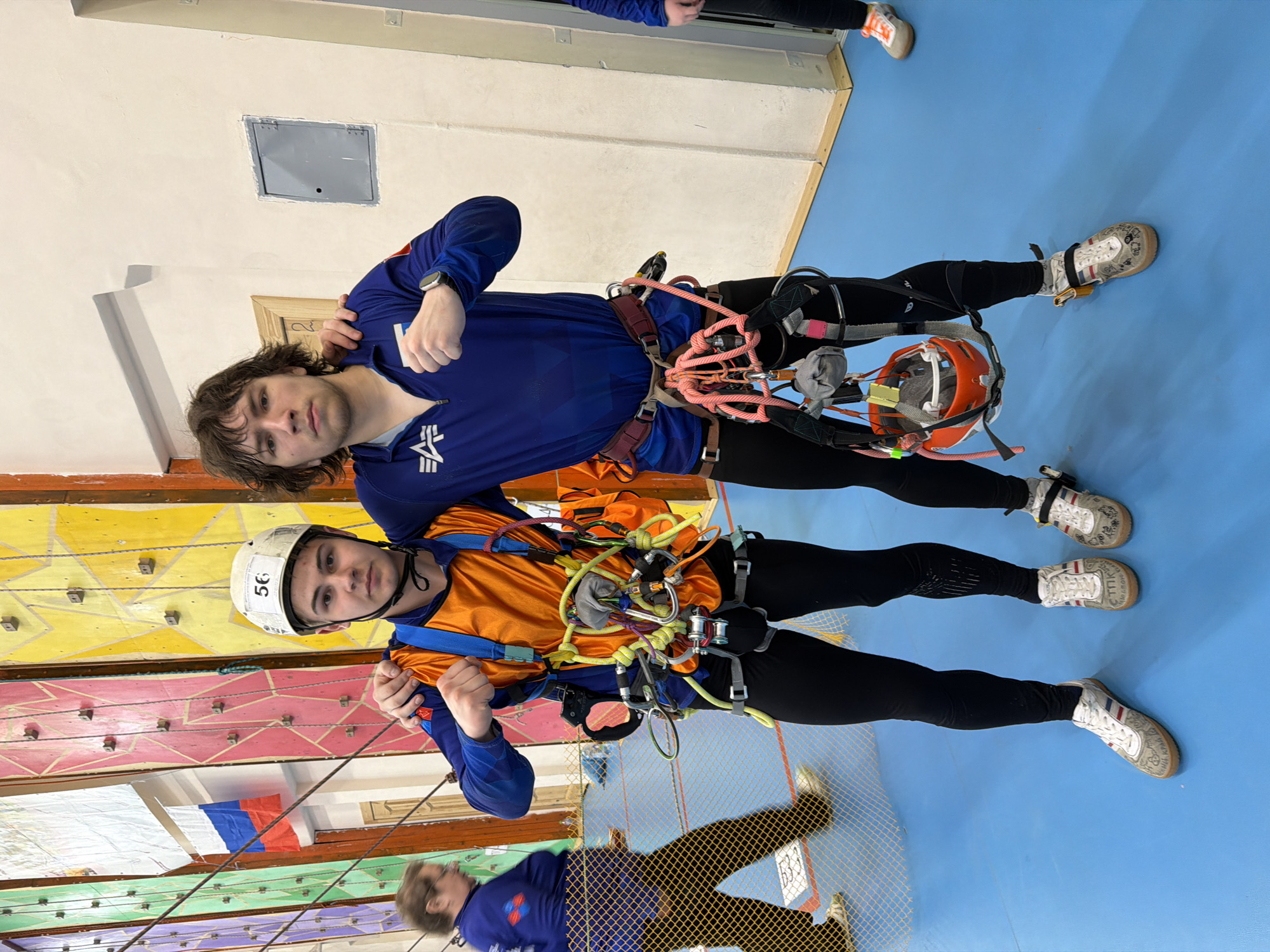 Two people are standing side-by-side, both wearing climbing harnesses and climbing shoes. The person on the left is wearing a blue long-sleeved shirt with an Alpha Industries logo on the left side, black pants, and a watch on their left wrist. The person on the right is wearing an orange and blue shirt, black pants, and a white climbing helmet with the number "56" on it. They are both indoors. Behind them is a climbing wall with various colored panels and holds. The background also shows a light blue floor and parts of other people in similar attire, indicating this is likely a climbing gym. The location is Berezniki, Russia.