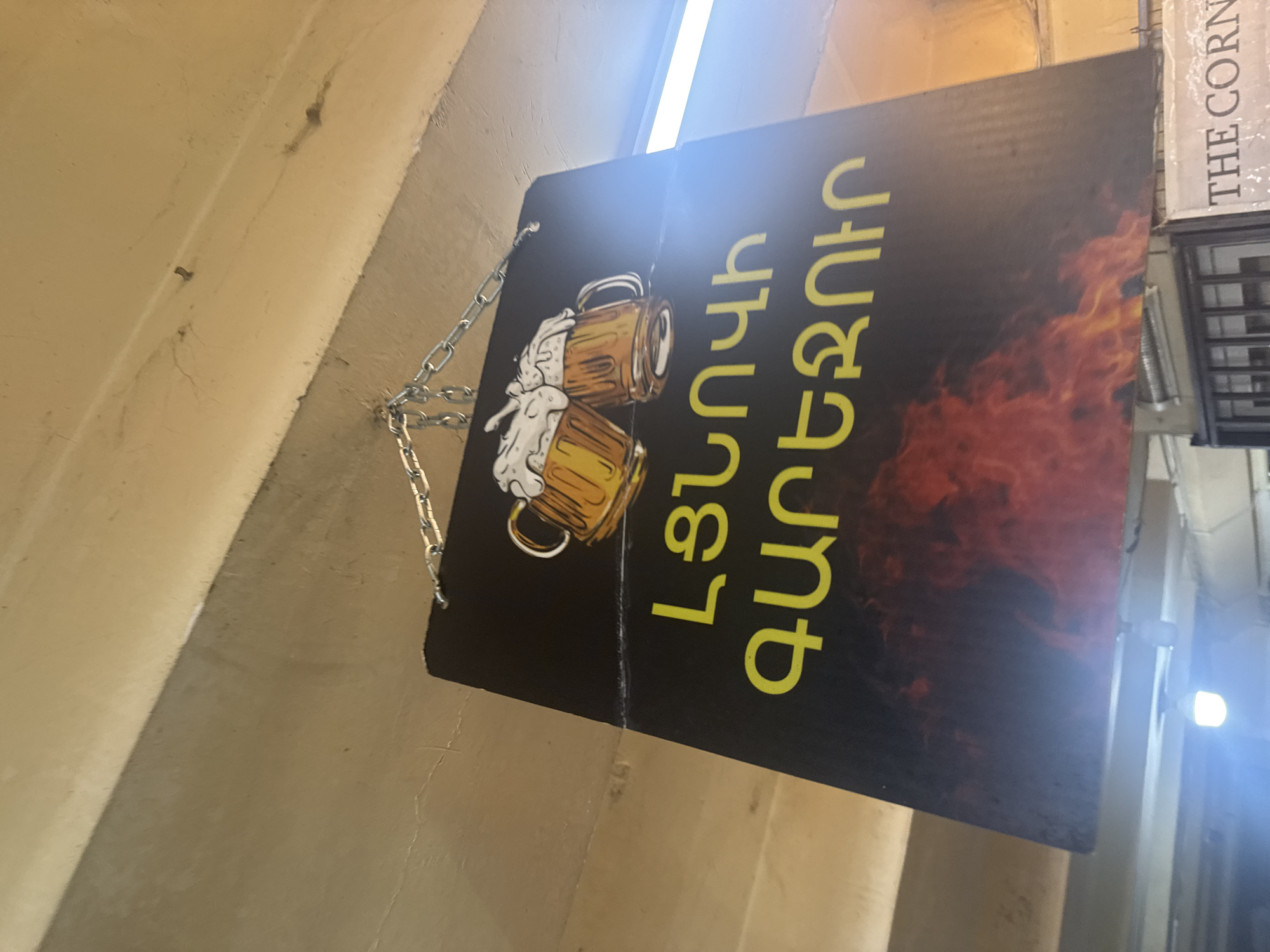 A sign hangs by chains from a building. The sign has a black background with two beer mugs and yellow Armenian writing. To the right of the sign is a vertical sign that says "The Corner." The sign is in Yerevan, Armenia.