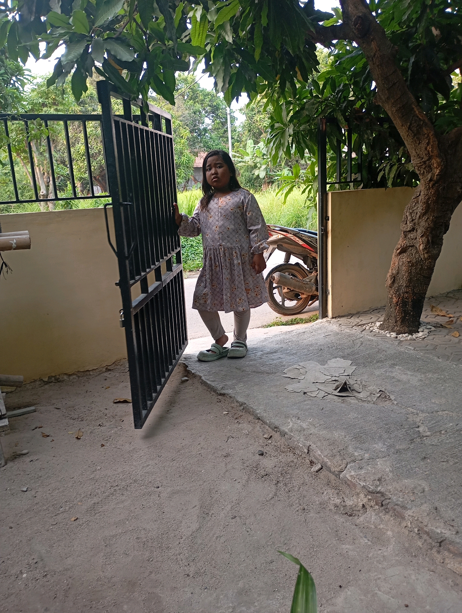 In Babelan, Indonesia, a young girl stands near an open black metal gate. She is wearing a long-sleeved, light-colored dress with a floral pattern and light-colored leggings, and appears to be looking towards the camera with a neutral expression. The gate is partially open, revealing a motorcycle behind it. The ground is a mix of concrete and dirt, with some debris scattered around. A large tree with rough bark is visible to the right of the gate, and lush green foliage forms a canopy overhead. The scene suggests a residential outdoor setting.