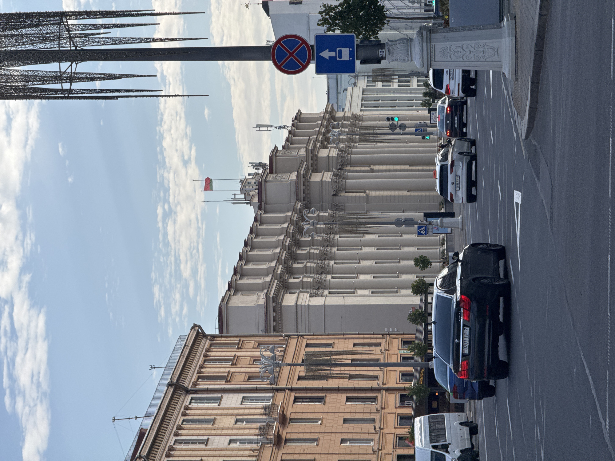 The image captures a street scene in Minsk, Belarus, featuring architectural elements and traffic. On the left, a tall, beige building with numerous windows and balconies lines the street. Adjacent to it, a grand, cream-colored structure showcases a long facade with a colonnade of classical columns and ornate detailing. A tall, modern sculpture, resembling icicles, stands prominently on the right side of the frame, its dark metallic structure contrasting with the sky.
The street itself is paved and marked with lane lines. Several cars are visible, including a black BMW sedan in the foreground with a license plate that appears to read "0701 TP-7". Other vehicles, including a white van and a white station wagon with a taxi sign on top, are also present. Traffic signals are visible, with one showing a green light.
Street signs are also present, including a "no stopping" sign and a directional sign indicating a bus route. The sky above is a pale blue, dotted with scattered white clouds. The overall impression is of a busy urban environment with a blend of historical and modern aesthetics. The specific location is likely near the Independence Square or a similar central avenue in Minsk, given the architectural style and the presence of governmental or significant public buildings implied by the grandeur of the structures.