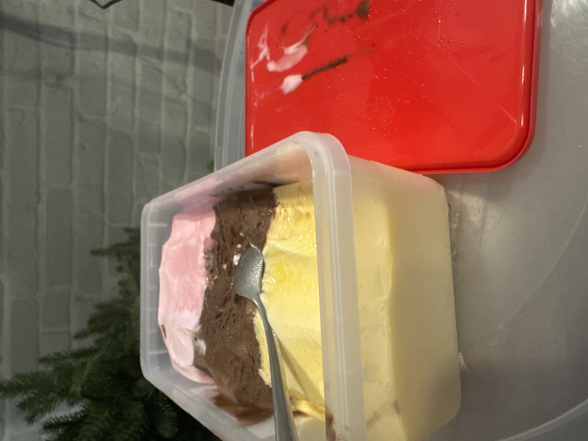 In an indoor setting, a transparent plastic tub filled with three distinct flavors of ice cream, arranged in horizontal layers, is positioned on a gray surface. The top layer is pink, the middle layer is brown, and the bottom layer is yellow. A silver spoon is partially submerged in the yellow ice cream. To the right of the tub, a bright red lid rests on the same gray surface. The background features a textured gray brick wall and a blurred green coniferous plant. The scene is static, with no visible activity or people. The lighting suggests an indoor environment. No specific location in Bangkok, Thailand can be determined from the visual information provided.