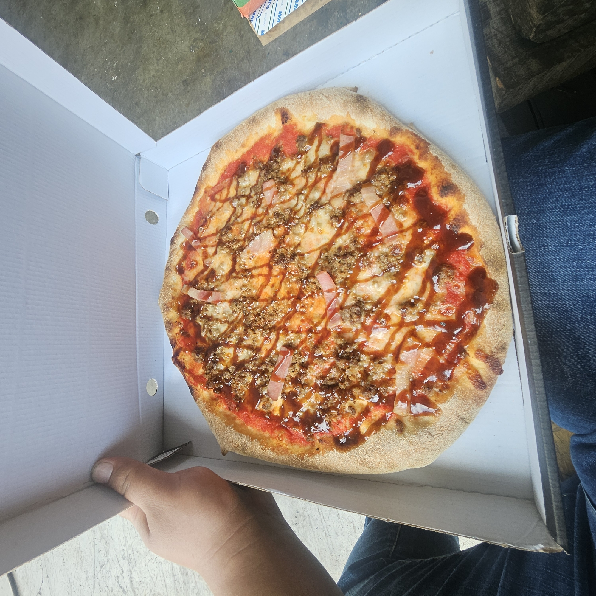 An overhead, medium shot shows a hand holding open a white pizza box containing a pizza. The pizza has a tomato sauce base, melted cheese, crumbled meat, and slices of ham, all drizzled with a dark, glossy sauce. The pizza is in a cardboard box that is resting on a person's lap, who is wearing blue jeans. The background appears to be an outdoor setting with a concrete floor. The hand holding the box is in the upper left quadrant of the image. The scene is likely in Mas Aliu, Spain, given the context.