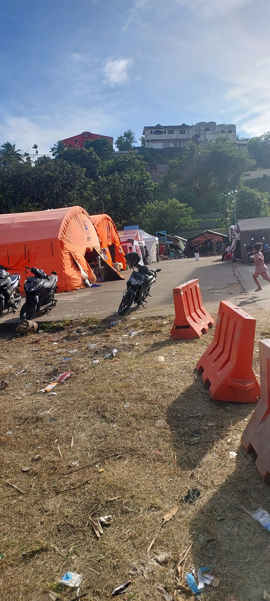 The image depicts an outdoor scene in Jayapura, Indonesia, likely a temporary settlement or aid distribution point. In the foreground, dry, sparse grass is littered with various pieces of trash, including plastic bottles and wrappers. Several orange, inflatable barriers are strategically placed in the foreground and midground, likely for traffic control or delineation of the area.
Behind the barriers, a paved surface is visible, on which several motorcycles are parked. Two large, orange tents, possibly emergency shelters or medical facilities, dominate the midground. One of these tents has "PKK" written on it. In front of these tents, smaller tents and structures can be seen, suggesting a more extensive setup.
Further back, several people are visible. A young child, wearing a pink garment, is captured in motion, running towards the right side of the frame. In the distance, on a hillside, several buildings are visible, including a distinctive red-roofed structure and a larger, multi-story building. The sky is blue with scattered clouds, and the overall lighting suggests it is daytime, possibly late afternoon due to the long shadows cast by the barriers. The environment appears somewhat rugged and undeveloped, consistent with a location in or near a city in Indonesia.