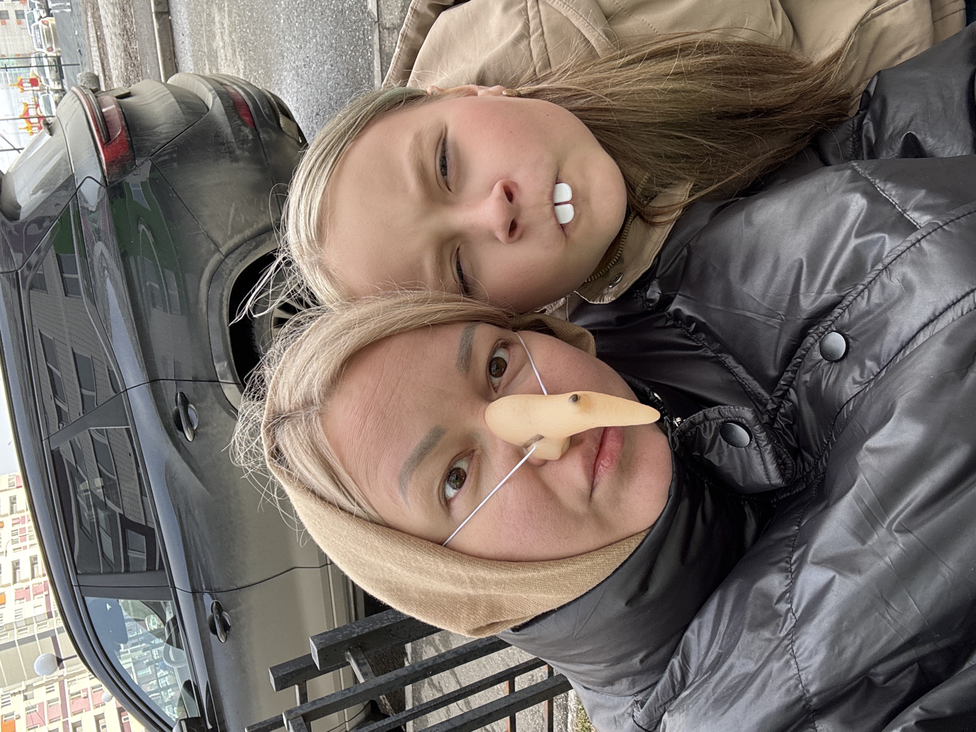 Two individuals, a woman and a younger girl, are photographed in a vertical close-up perspective. The woman is wearing a black puffer jacket and what appears to be a tan hood or scarf. She also has an artificial long nose attached with a thin white string. The girl, who is lying next to her, is wearing a tan jacket and has what appear to be two white objects inserted in her mouth, possibly simulating teeth. A black car is visible in the background, slightly out of focus. The location, as stated in the prompt, is Izhevsk, Russia. The environment appears to be urban, possibly near a street or parking area, as indicated by the car and a metal fence.