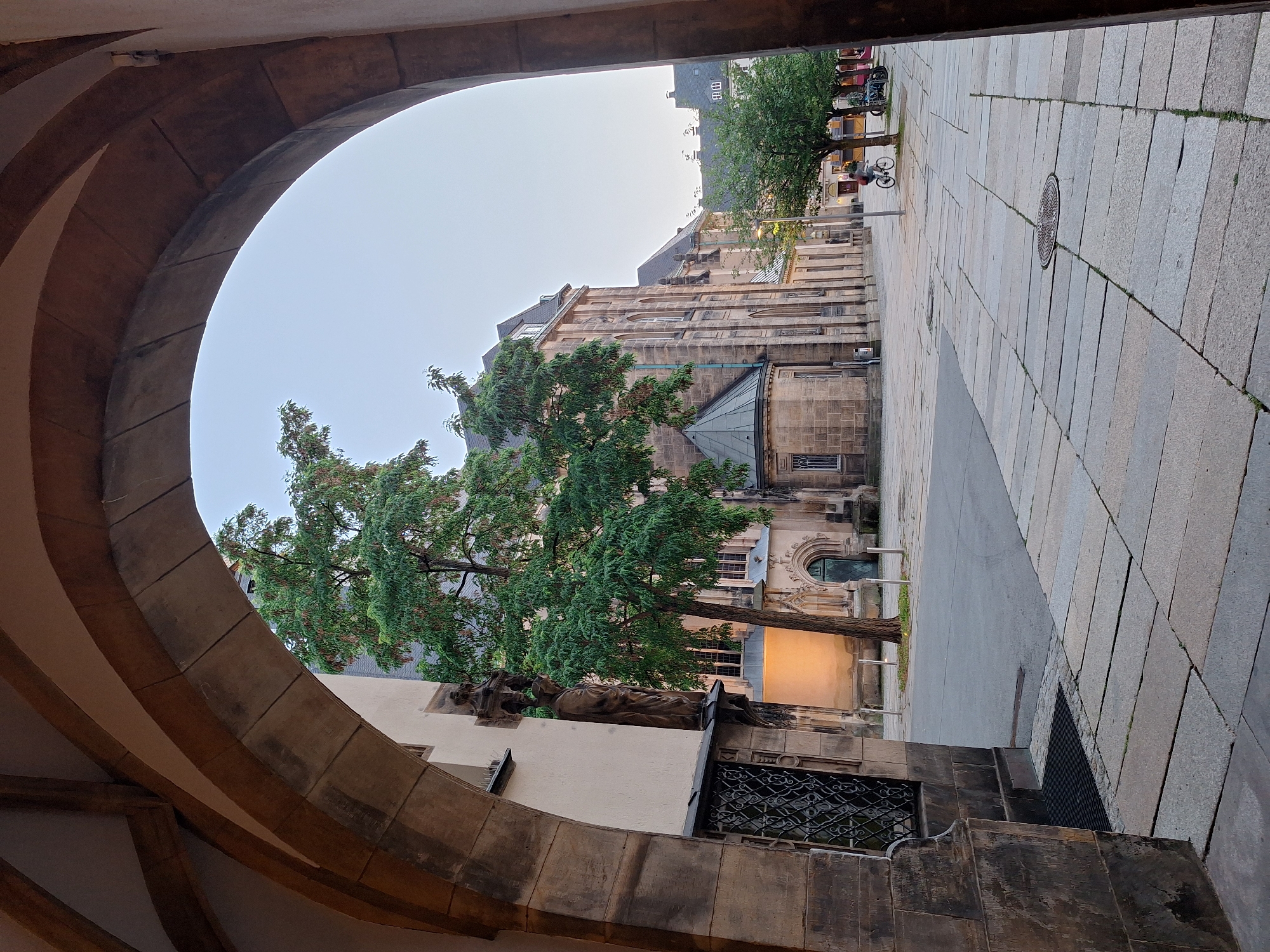 The image captures an outdoor scene framed by a stone archway. Beyond the arch, a street lined with stone buildings is visible. In the foreground, the stone archway creates a sense of depth and perspective. The street is paved with large, rectangular stone tiles, and a manhole cover is visible on the pavement.
To the left, a building with stone facade and arched windows is partially obscured by a large tree with green foliage. The tree's branches extend across the scene, some appearing reddish or pink, possibly indicating flowers or fruits. A statue or sculpture of a reclining figure can be seen on the building's exterior, near the base of the tree. To the right of the street, another building with a grey roof and stone construction is visible, with multiple windows and architectural details.
In the background, more buildings and trees can be seen. There are several bicycles parked along the street, and a person is riding a bicycle down the street, moving away from the viewer. The overall lighting suggests it is daytime, possibly late afternoon or early morning given the soft light. The sky is a pale, overcast grey. The scene appears to be in a historical or European city center, given the architectural style and cobblestone paving. The environment seems calm and serene. No discernible text is present in the image.