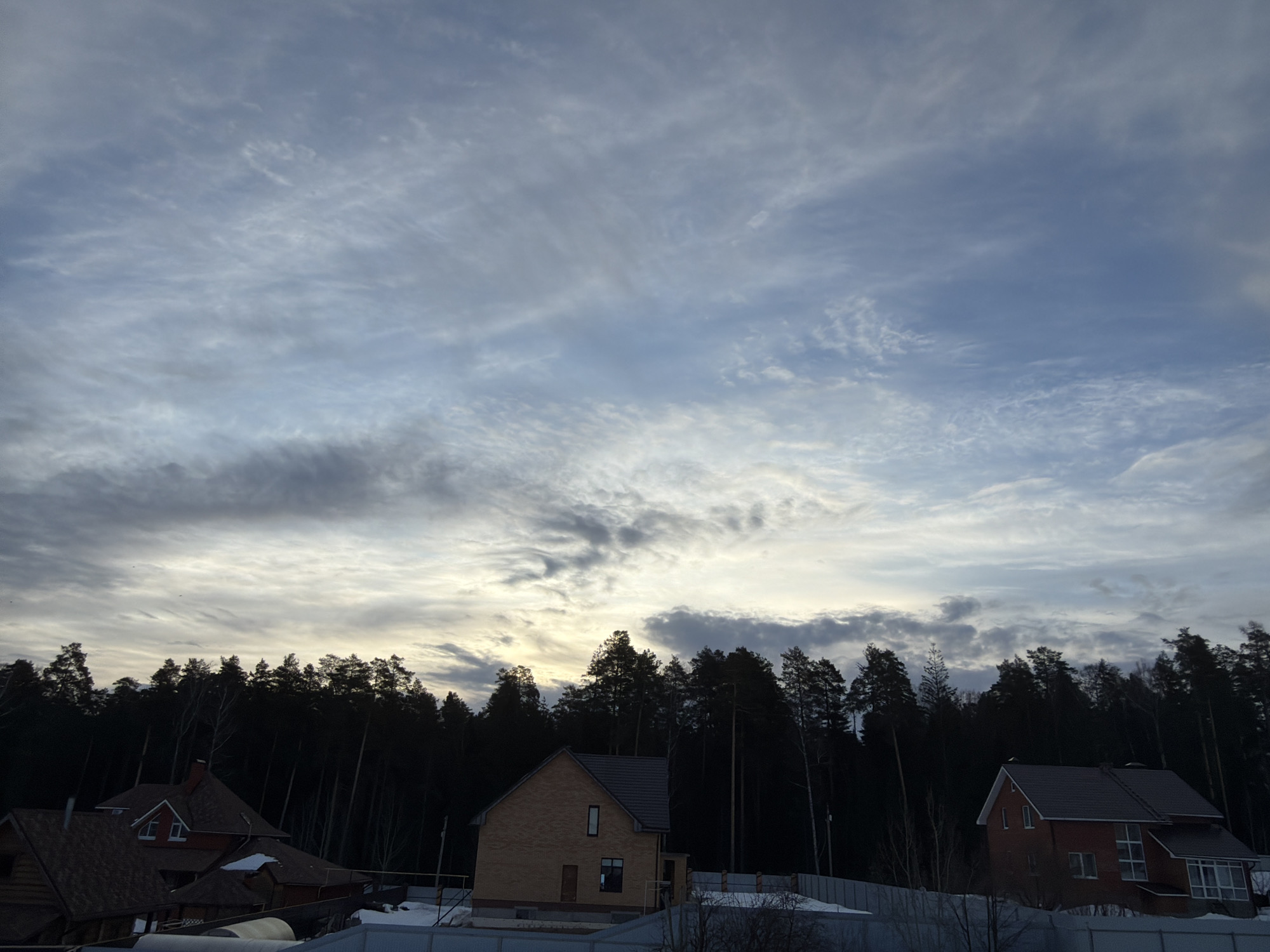 The image displays a residential area in Chaikovsky, Russia, under an overcast sky. The upper two-thirds of the frame are dominated by a sky featuring numerous cirrocumulus and stratocumulus clouds, exhibiting varying shades of grey, white, and pale blue, with a subtle yellowish-white luminescence near the horizon. A dense, dark coniferous forest line stretches across the midground.
In the lower third of the image, three residential structures are visible, with patches of snow on the ground indicating a cold season. On the far left, a portion of a brown wooden house with a reddish-brown tiled roof is present. Centrally, a two-story, light tan brick house with a dark grey sloped roof stands, featuring a single vertical window on the upper front facade and a door with a window on the ground level. To the right, a two-story red brick house with a dark grey sloped roof is visible, characterized by multiple windows, including a large multi-pane window on its ground floor. A light-colored corrugated fence runs across the foreground, partially obscuring the bases of the properties. No individuals or vehicles are present.