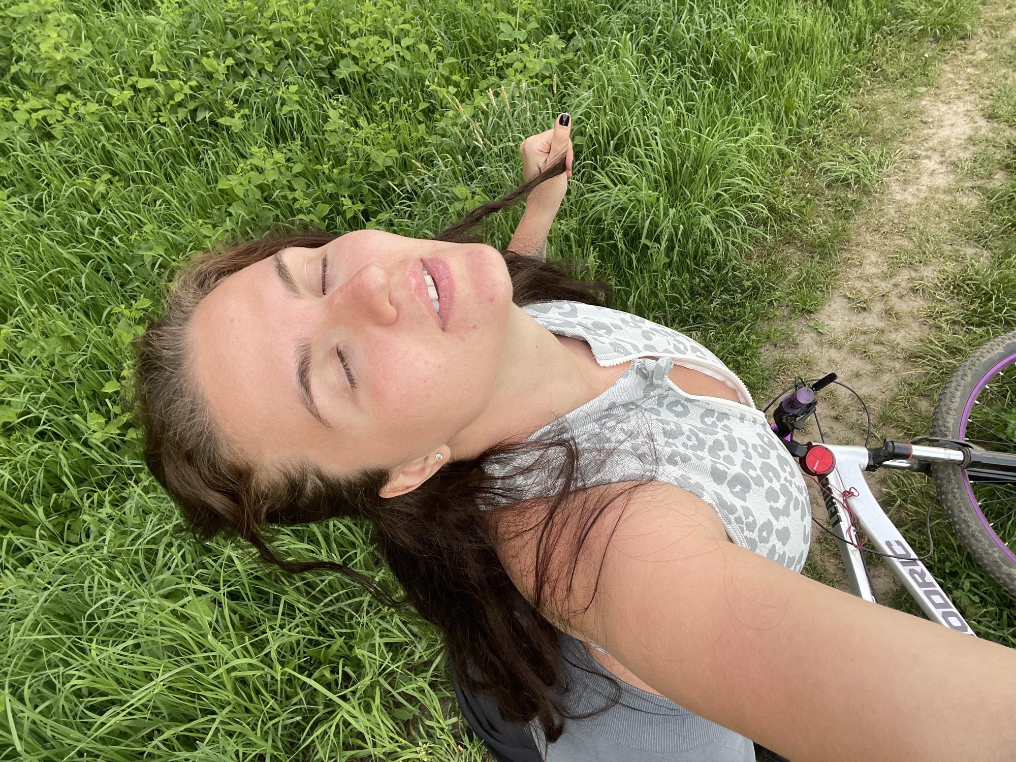 A young woman is lying on her back in a grassy field, holding a strand of her dark brown hair in her right hand. She is fair-skinned with dark hair and appears to be smiling with her eyes closed. She is wearing a gray and white animal print top. A bicycle with purple rims is visible next to her, positioned on a dirt path adjacent to the grass. The location cannot be confirmed as Ostrov, Russia without further information. The environment consists primarily of grass and a dirt path.