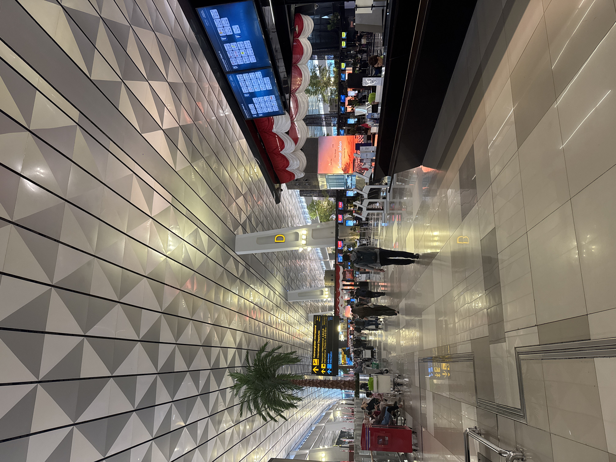 The image captures an interior view of an airport terminal, likely Soekarno-Hatta International Airport in Tangerang, Banten, Indonesia, near the city of Pajang. The terminal features a modern, expansive design with a geometrically patterned ceiling. Several passengers are seen walking through the concourse, some with luggage. A prominent check-in area is visible, complete with digital departure boards displaying flight information. Decorative red and white drapery, possibly reflecting Indonesian national colors, hangs above the check-in counters. A tall artificial palm tree adds a tropical element to the indoor environment. The floor is polished and reflects the overhead lighting and architectural features.