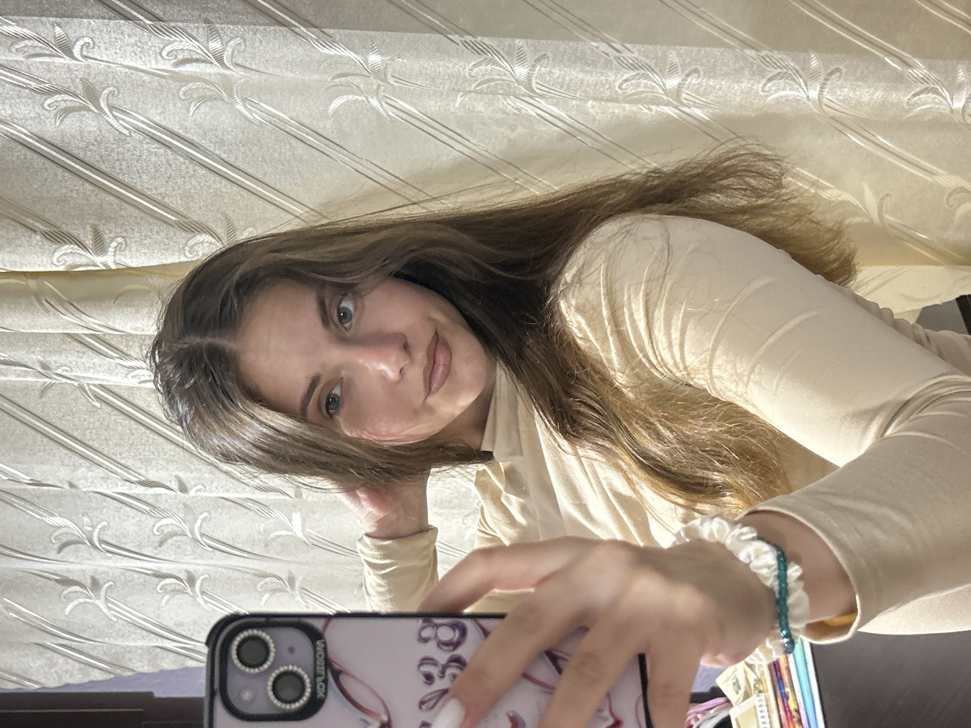 An image shows a young woman with long, brown hair taking a selfie in front of a cream-colored, patterned curtain. She is wearing a light beige, long-sleeved shirt. Her left hand is raised to her head, holding her hair, and her right hand holds a smartphone with a patterned case, displaying what appears to be a pink and purple design and the number "38". She is wearing a white scrunchie with a beaded bracelet on her left wrist. The lighting suggests it is indoors, possibly in a bedroom or living area, with the light source casting shadows on the curtain. The background curtain has a repeating floral or leaf-like pattern with diagonal lines.