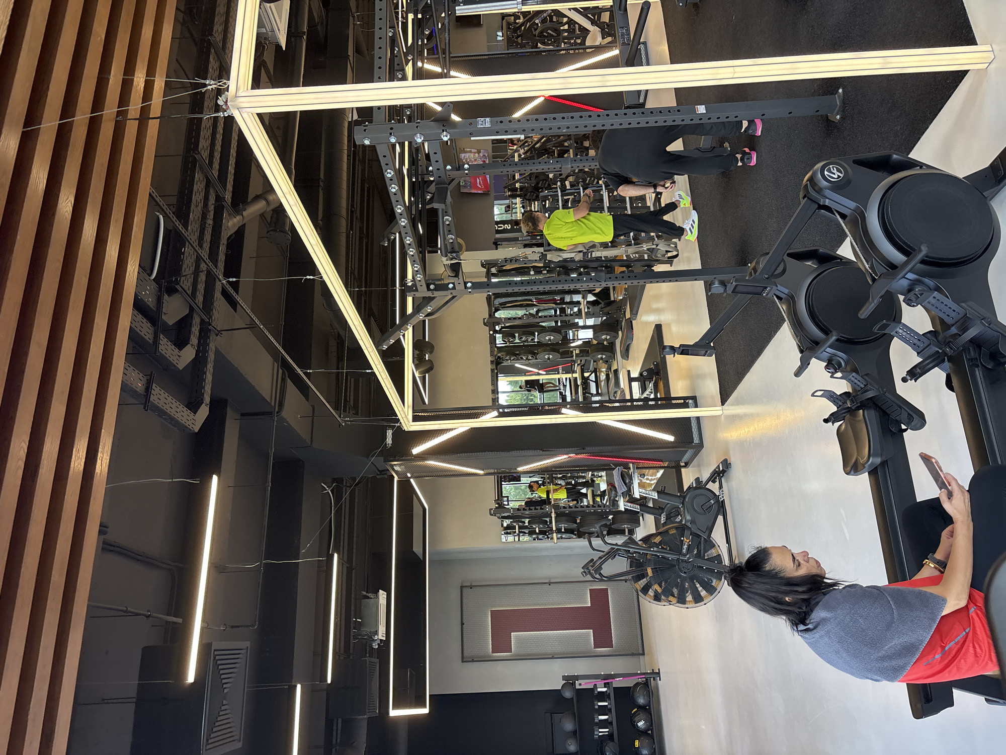 The image depicts the interior of a modern gym. In the foreground, a woman with dark hair tied back in a ponytail, wearing a red t-shirt and a grey towel draped over her shoulders, is seated on a rowing machine and looking at her phone. She is positioned on the left side of the frame. Behind her and to the right, two individuals are visible. One person, wearing a bright yellow shirt and black pants, is standing near weight racks. Another person, wearing black attire, is bent over near the weight racks. The gym is equipped with various exercise machines, including rowing machines, air bikes, and weightlifting equipment, such as squat racks and dumbbell racks. The environment features industrial-style lighting, a black ceiling with exposed conduits, and light-colored flooring. A large graphic of the number "1" is prominently displayed on a wall in the background, to the left of the frame. The overall scene suggests a functional and contemporary fitness facility. The location is identified as Monolit, Russia.